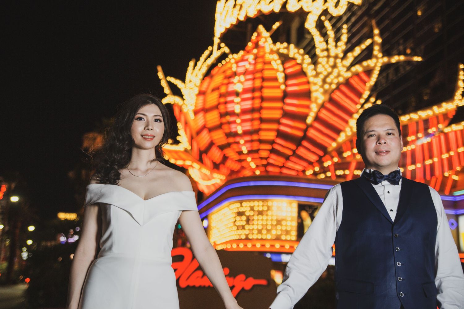 Couple holds hands in front of glowing Flamingo casino sign at night in Las Vegas.