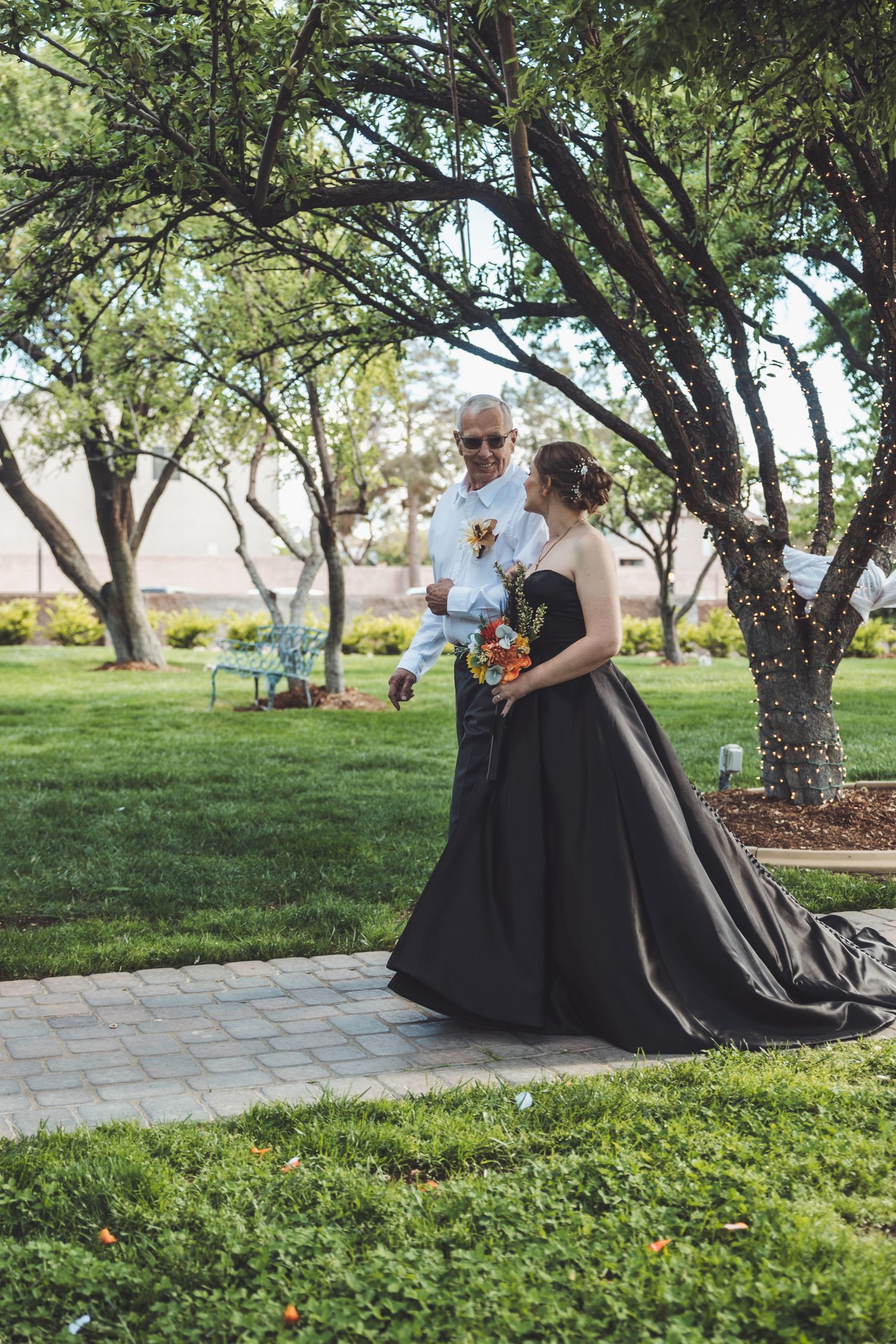 Bride in black wedding gown walks with older man through garden on stone path surrounded by green trees.