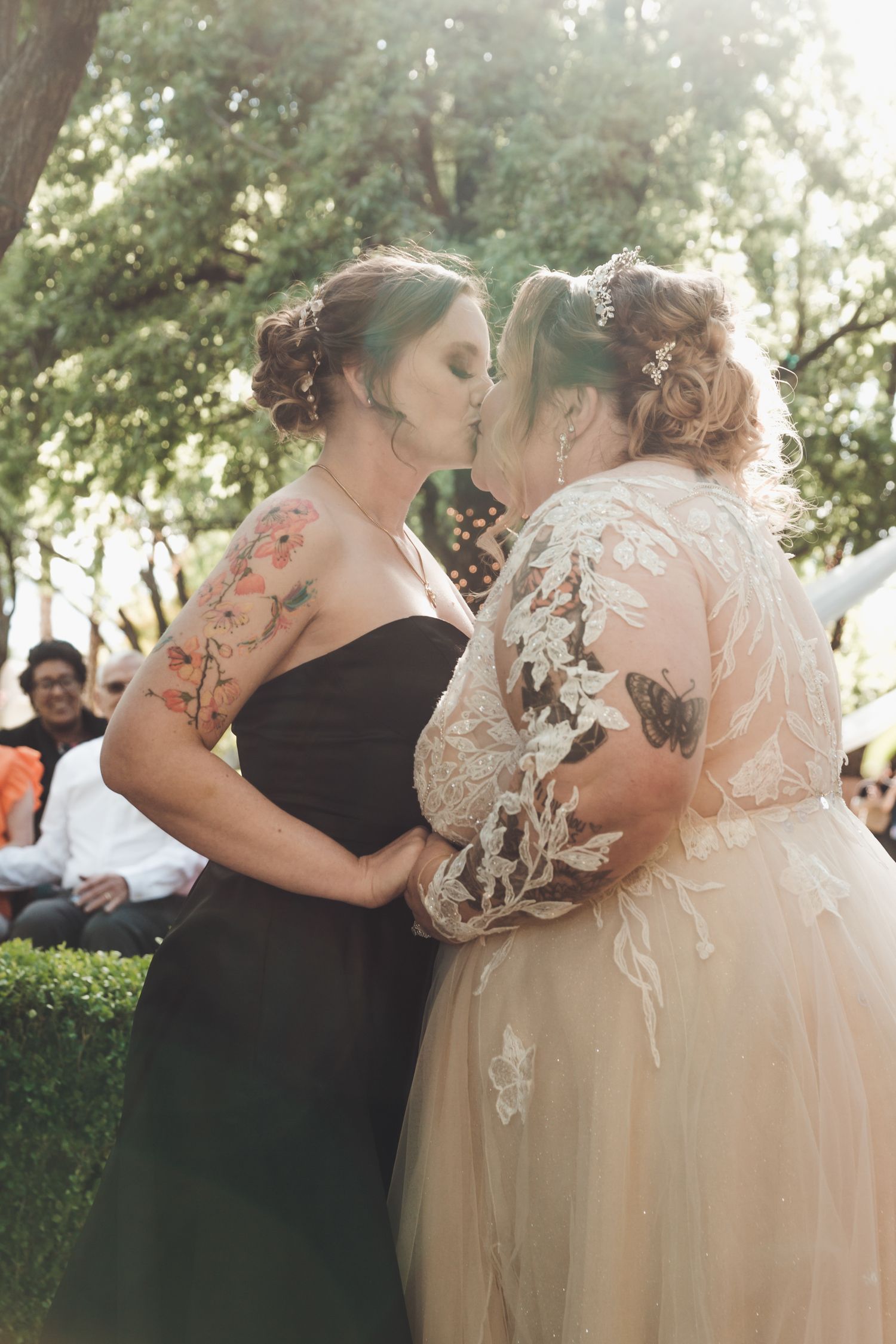 Two brides share first kiss during outdoor wedding ceremony, one in black dress, one in lace gown.