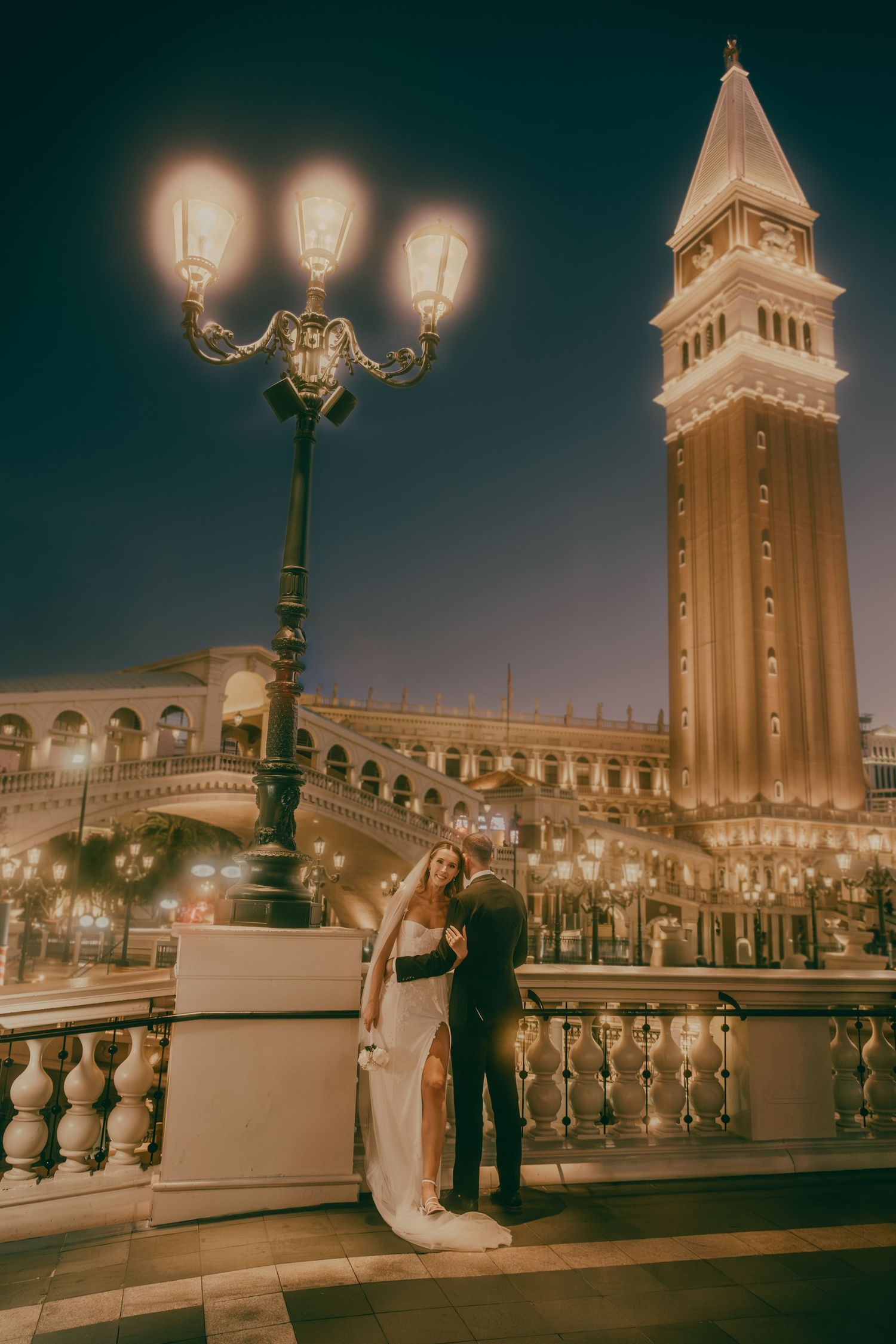 Wedding couple embrace near an ornate lamppost with the Venetian tower illuminated against the night sky.