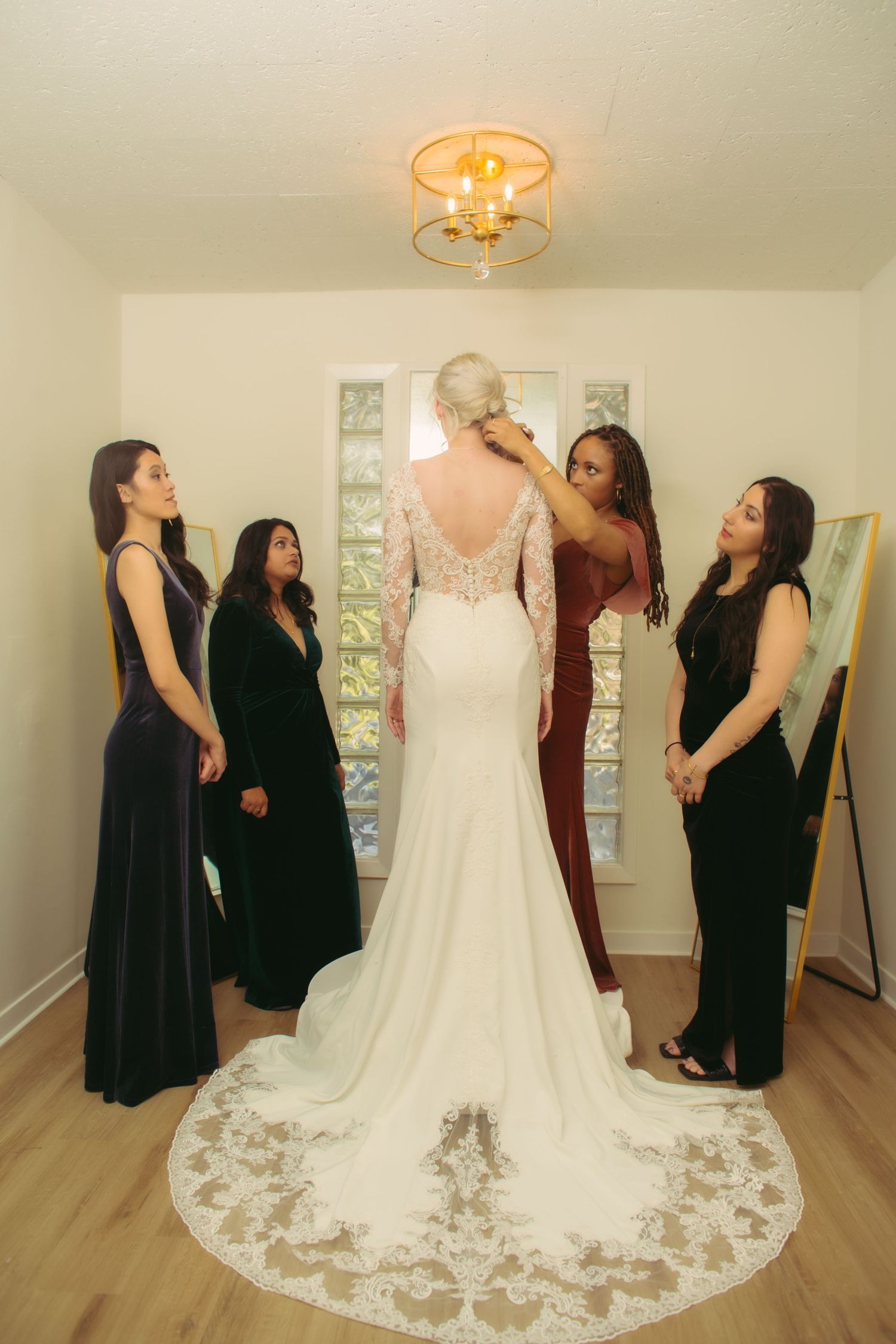 Four bridesmaids surrounding bride in lace-back mermaid wedding gown with cathedral train indoors.