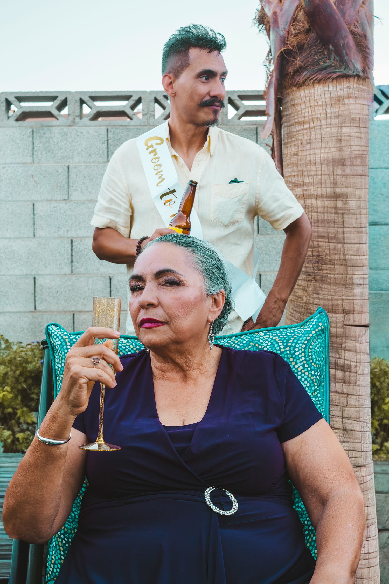 Older woman in navy dress holds champagne glass while man in white shirt with sash stands behind her outdoors.