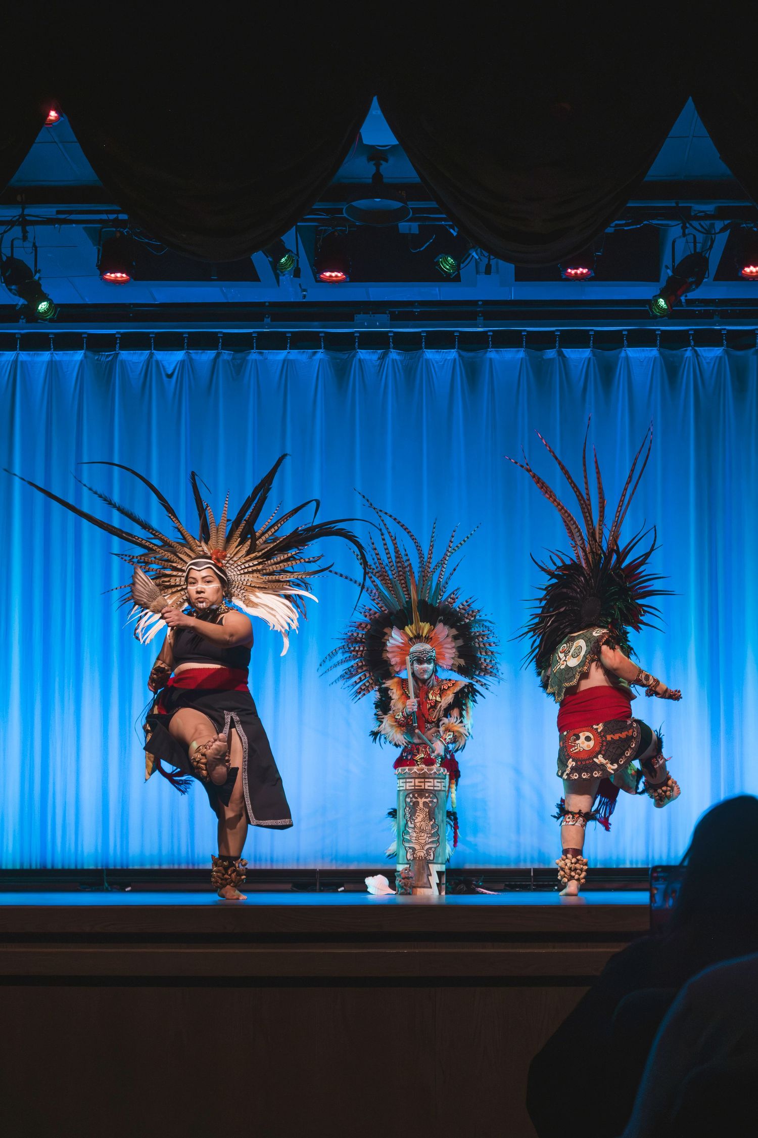Three performers in traditional Aztec headdresses and costumes dancing on a blue-lit stage.