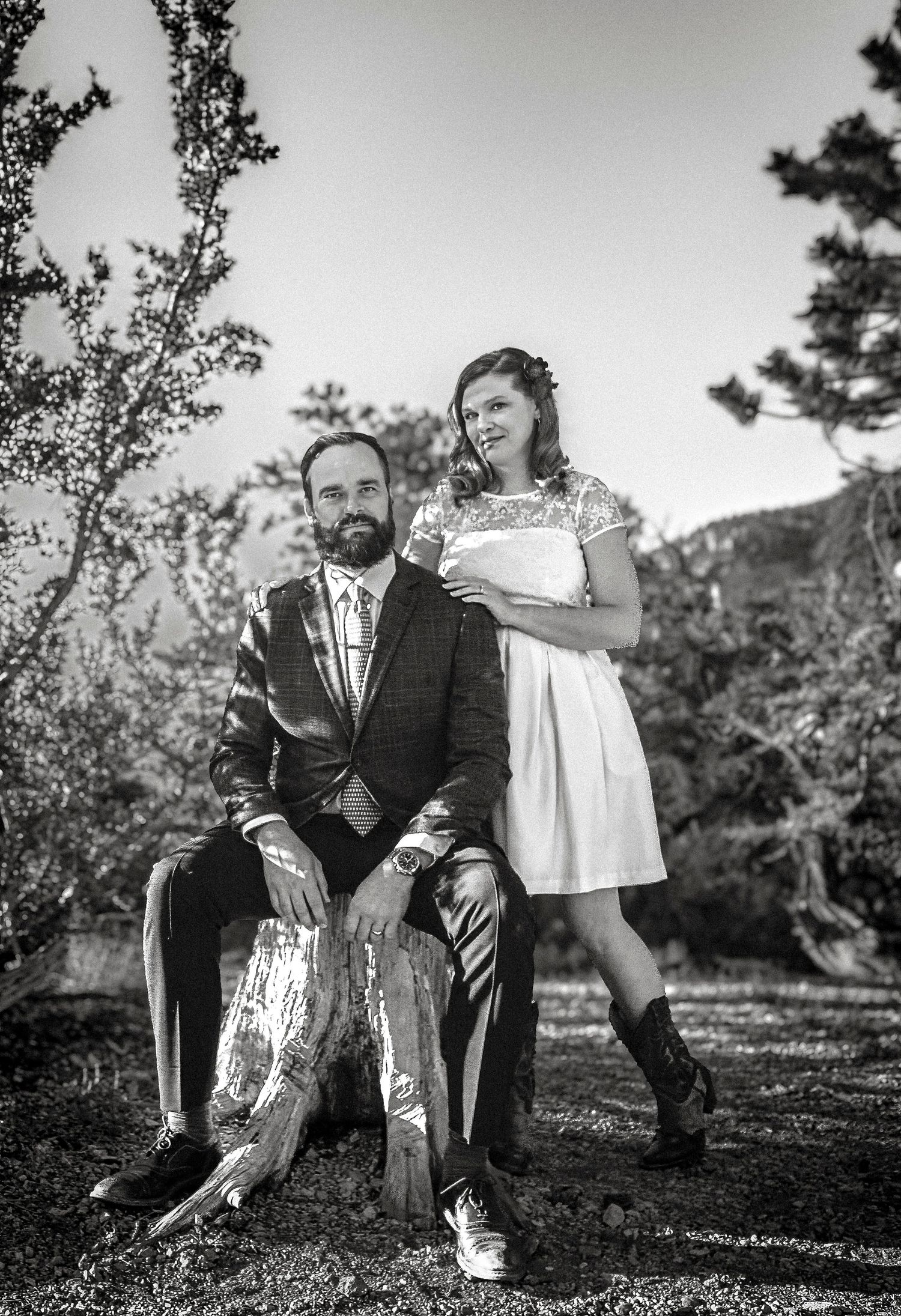 Black and white wedding photo of a bearded groom seated and bride standing beside him outdoors.
