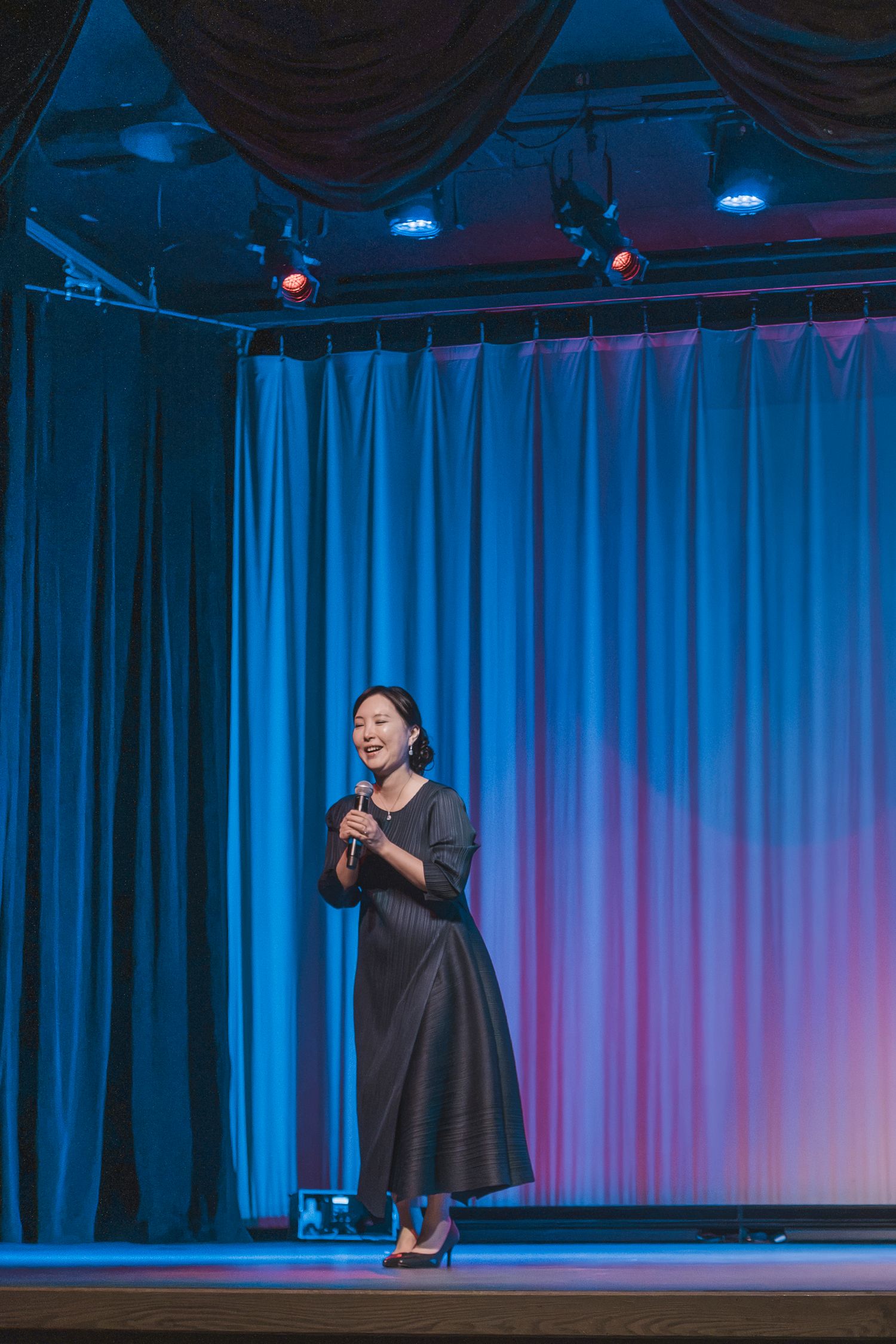 Woman in gray dress speaking on stage with blue and pink curtain backdrop under stage lights.