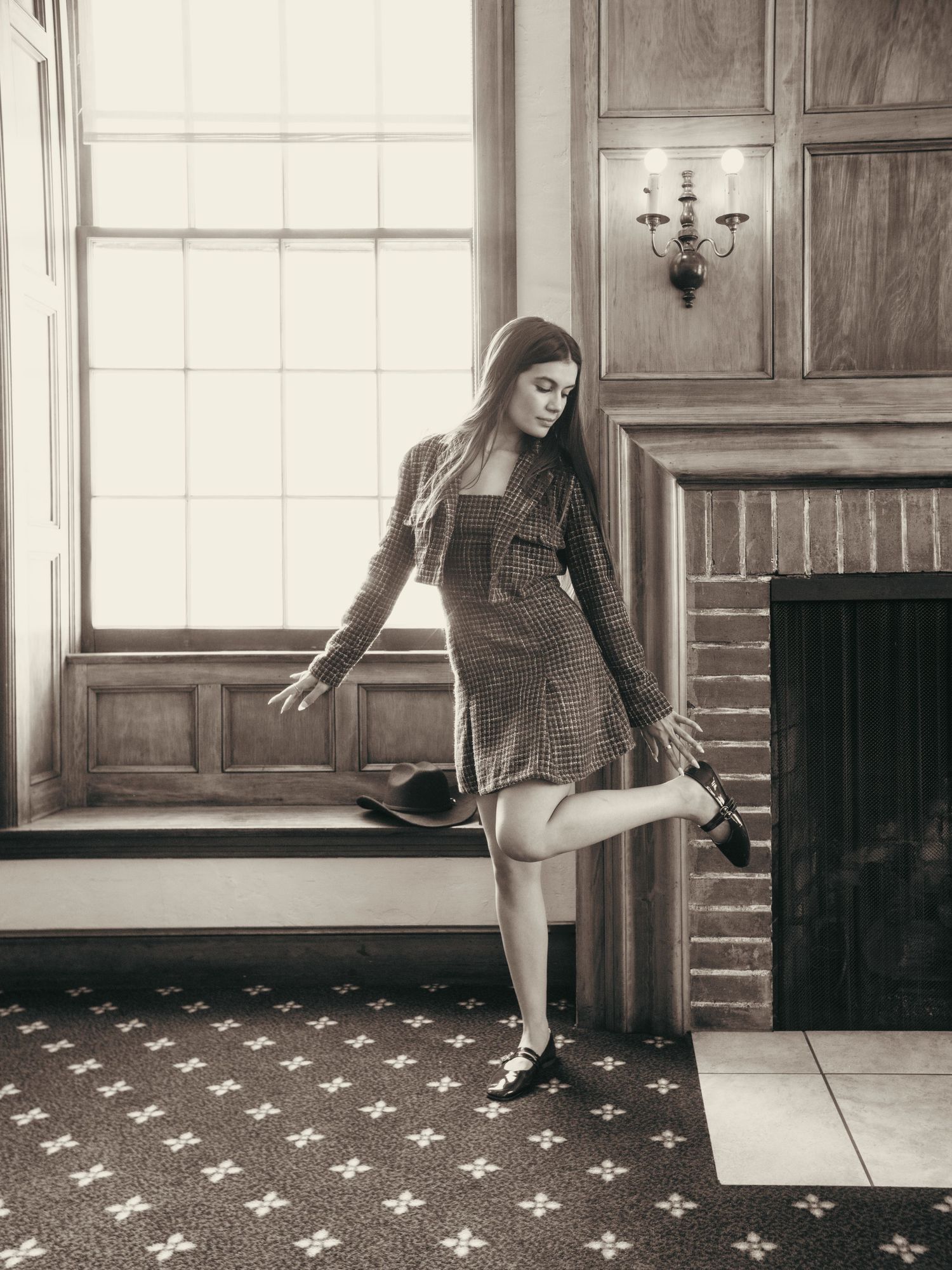 Young woman in plaid dress poses playfully by vintage window near brick fireplace in sepia-toned interior photo.