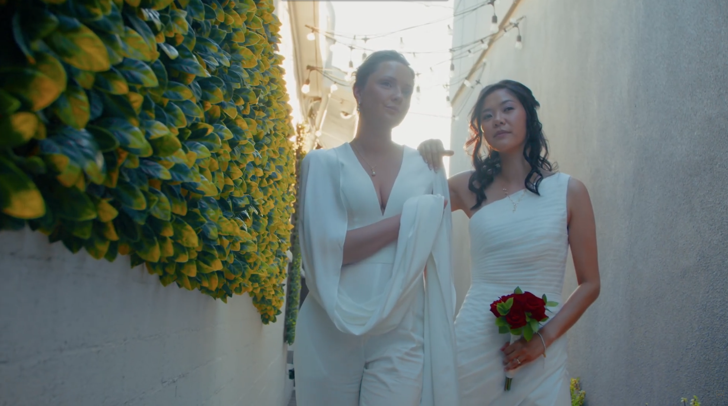 Two brides in white gowns stand in a narrow alley adorned with yellow flowers, one holding a red bouquet.