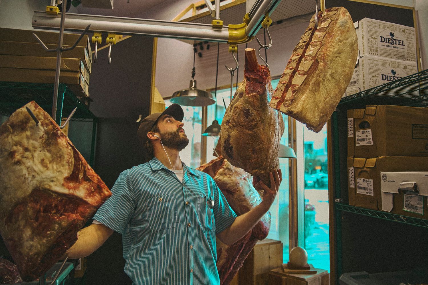 Butcher inspecting hanging dry-aged meat cuts in a dimly lit meat locker storage room.
