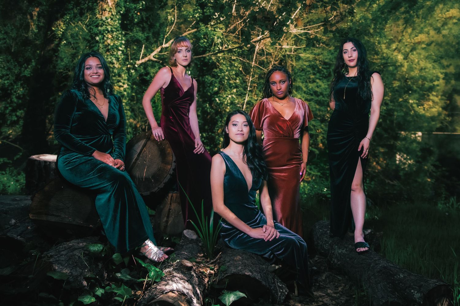 Five women in elegant velvet gowns pose gracefully in a lush forest setting for a glamorous outdoor photo shoot.