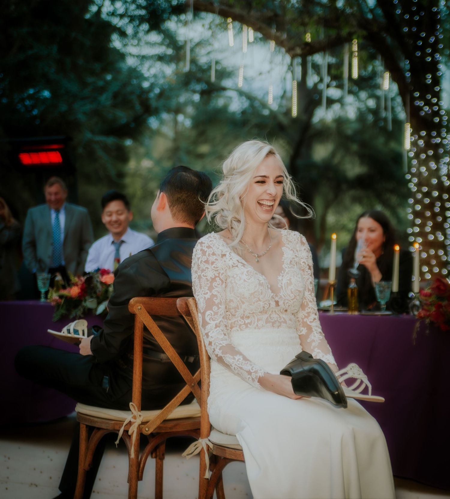 Bride in lace gown laughing joyfully at outdoor wedding reception with guests in background.