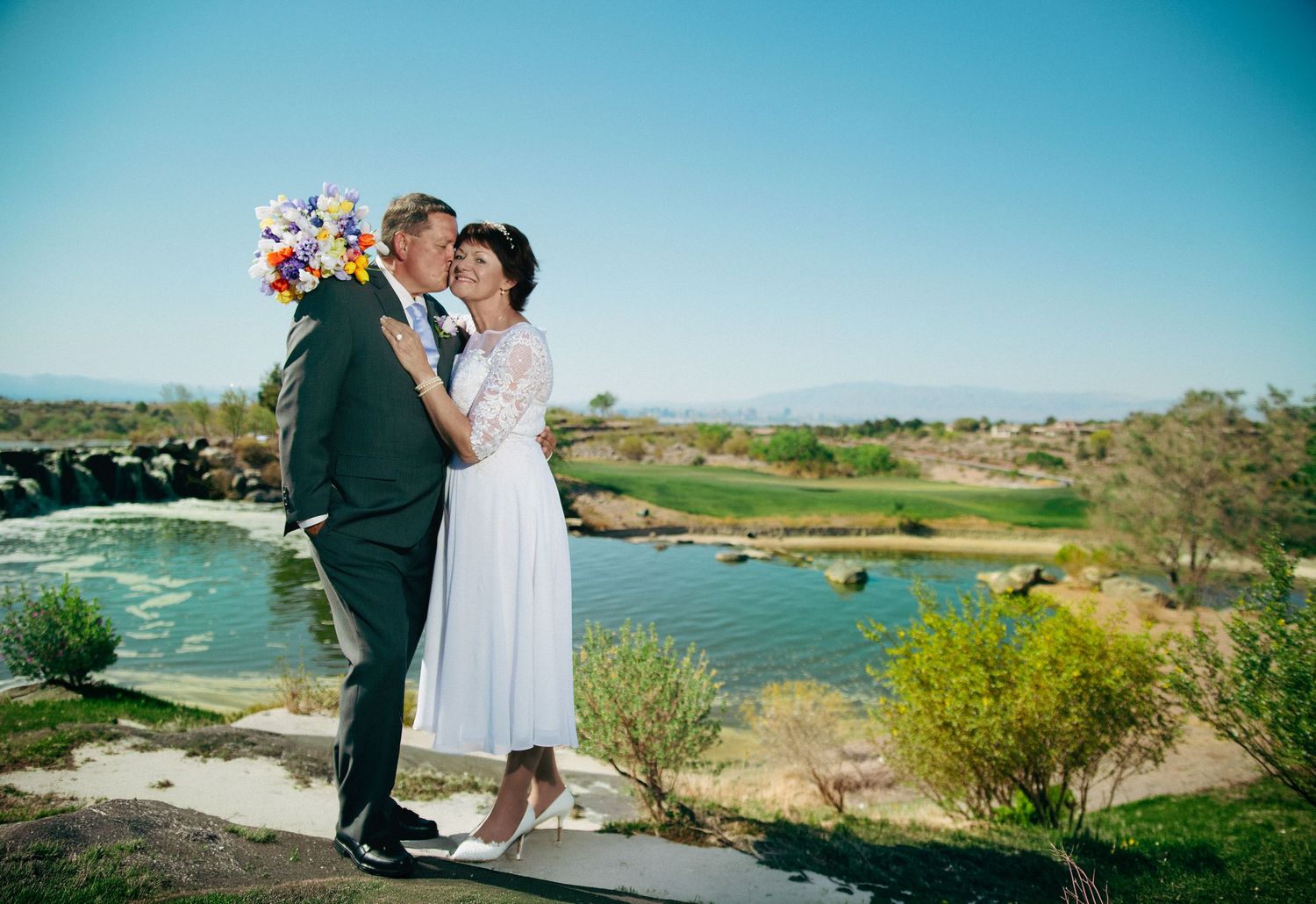Bride and groom share a kiss outdoors near a scenic lake on their wedding day, bride holding colorful bouquet.