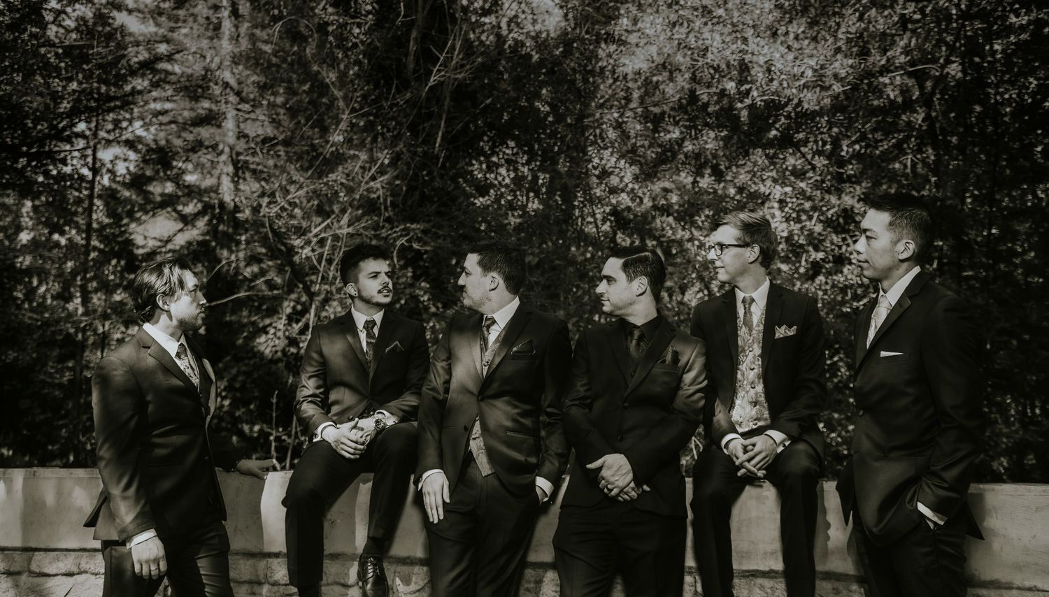 Six groomsmen in dark suits laughing and socializing outdoors in a black and white wedding photo.