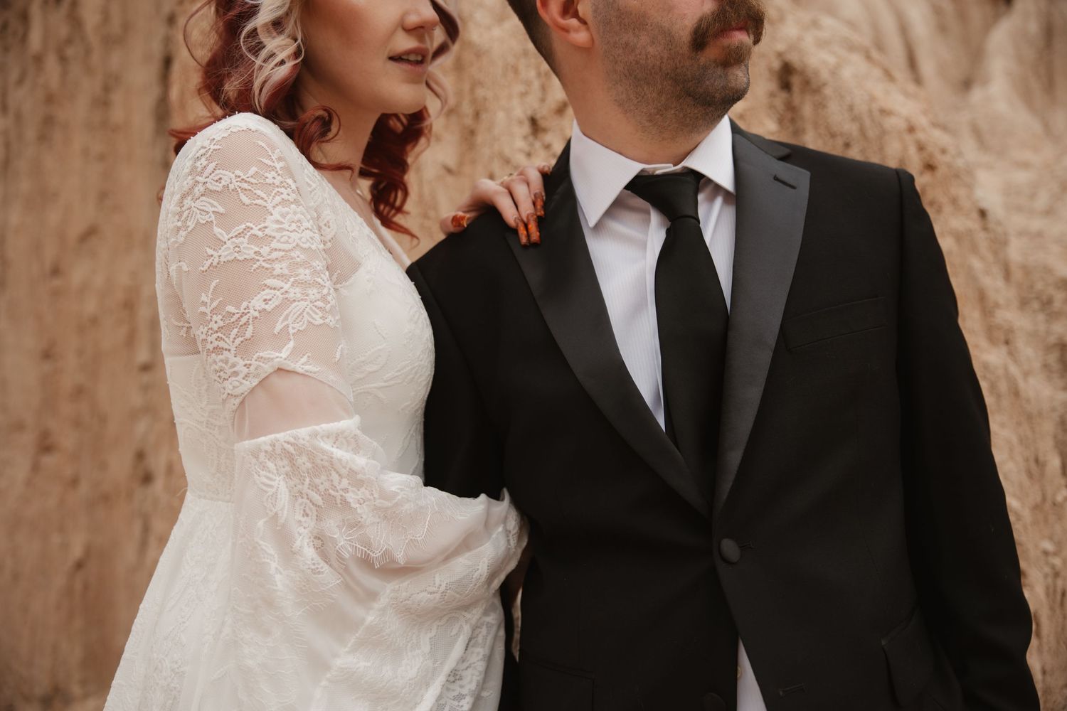 Wedding couple poses together, bride in white lace gown and groom in black suit with tie outdoors.