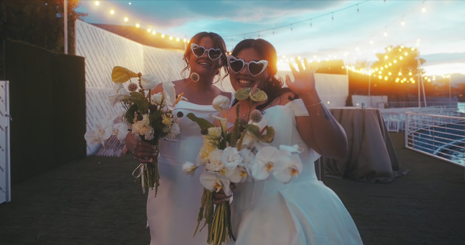 Two brides in white dresses wearing sunglasses holding bouquets at sunset.