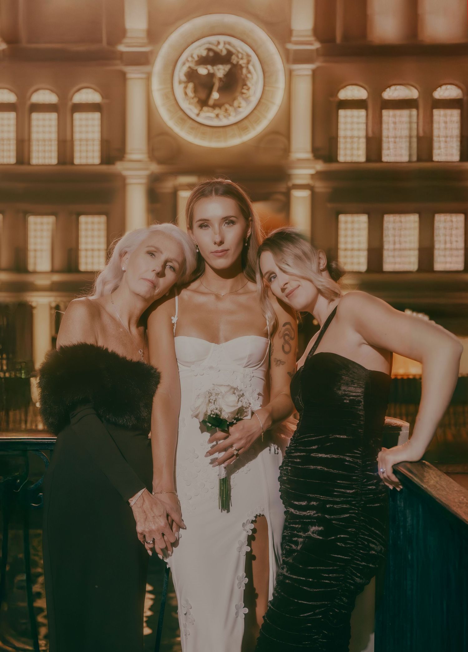 Bride and two guests in black dresses pose seriously before grand illuminated clock at Las Vegas venue.