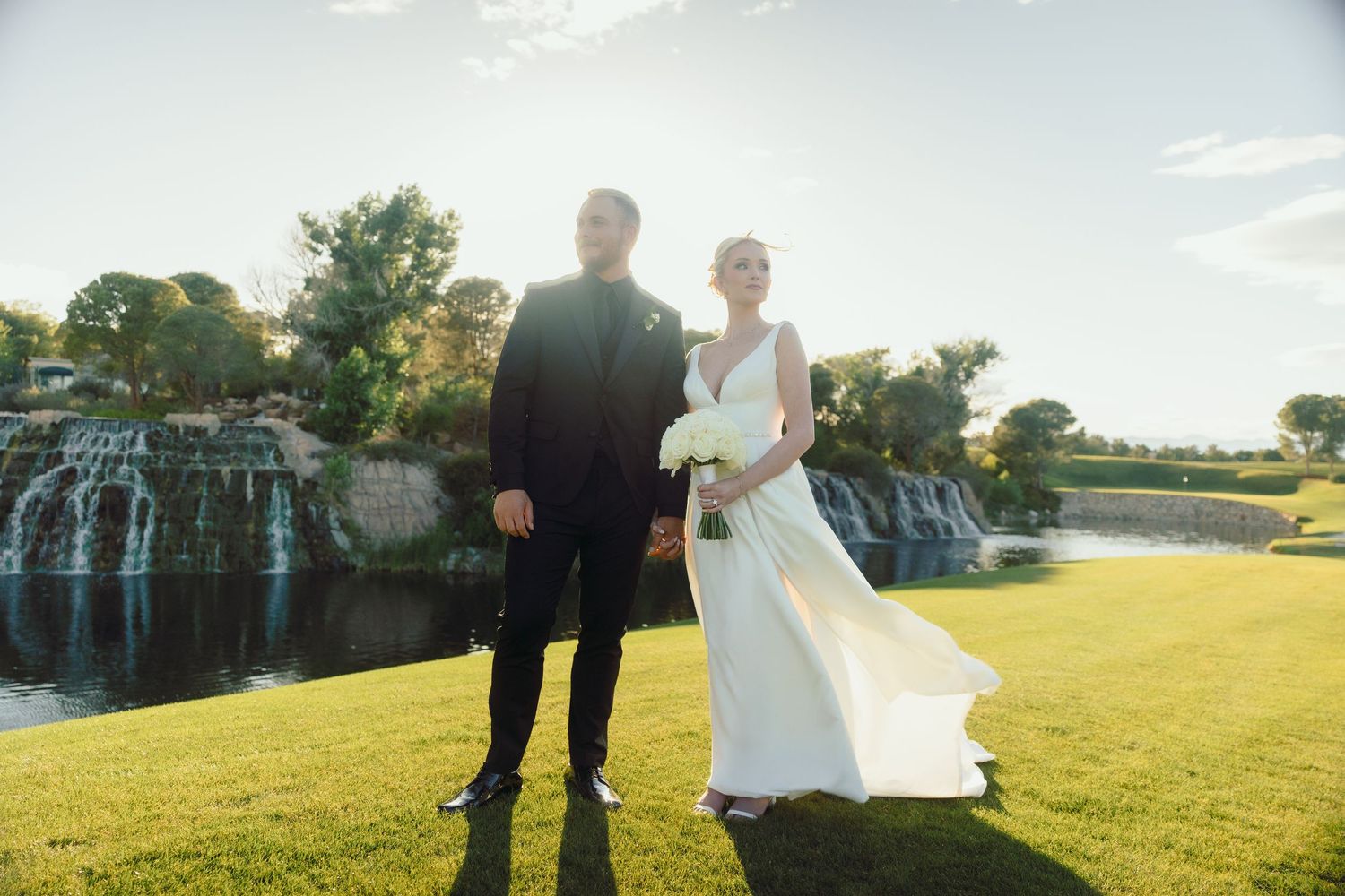Bride and groom standing on green lawn with waterfall backdrop in sunlight.