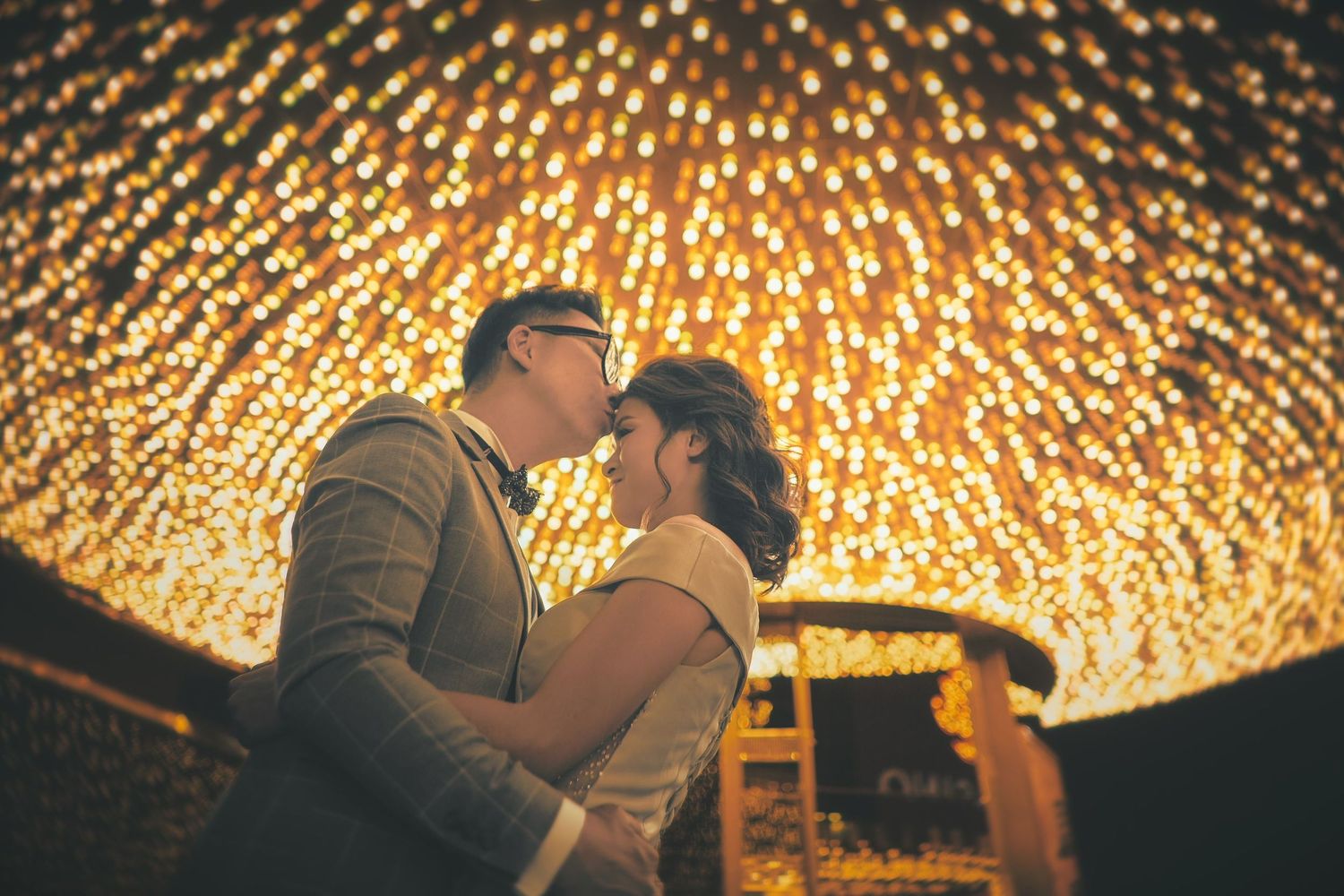 Couple sharing a romantic kiss under a canopy of warm golden bokeh lights at night.