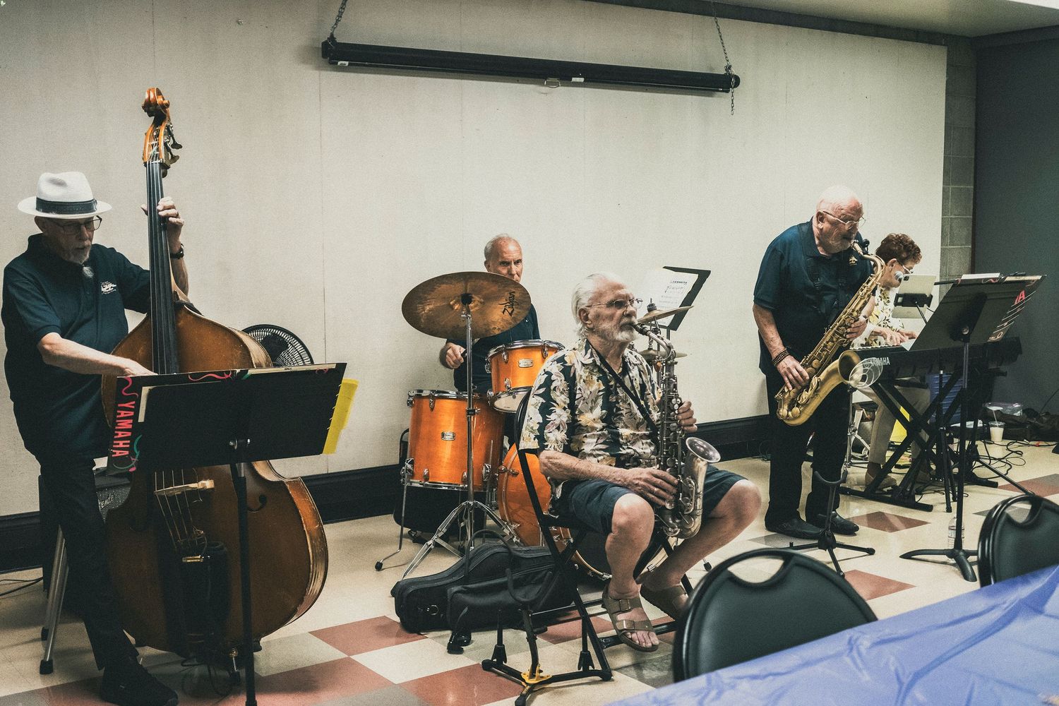Jazz band performing indoors with upright bass, drums, and saxophone players in a casual rehearsal space.