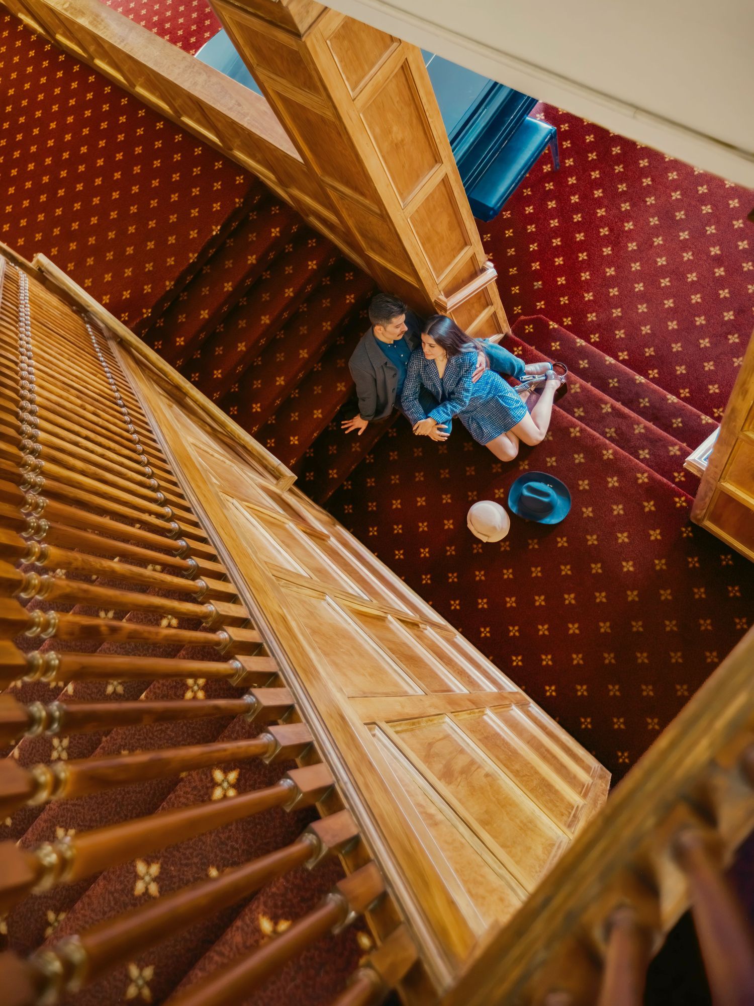 Aerial view of couple sitting on red carpeted grand staircase with wooden banisters in elegant hotel.