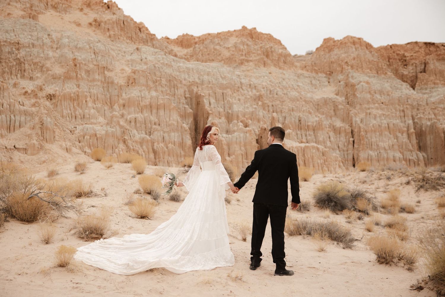 Bride in flowing lace gown and groom in black suit hold hands before stunning red desert cliffs.