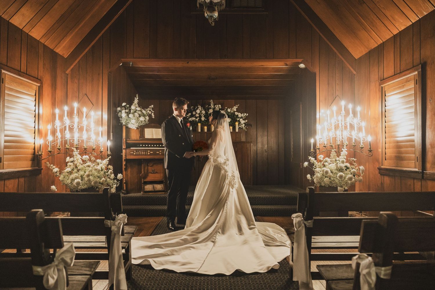 Bride and groom share intimate moment at candlelit wooden chapel altar with floral arrangements and elegant trailing veil.