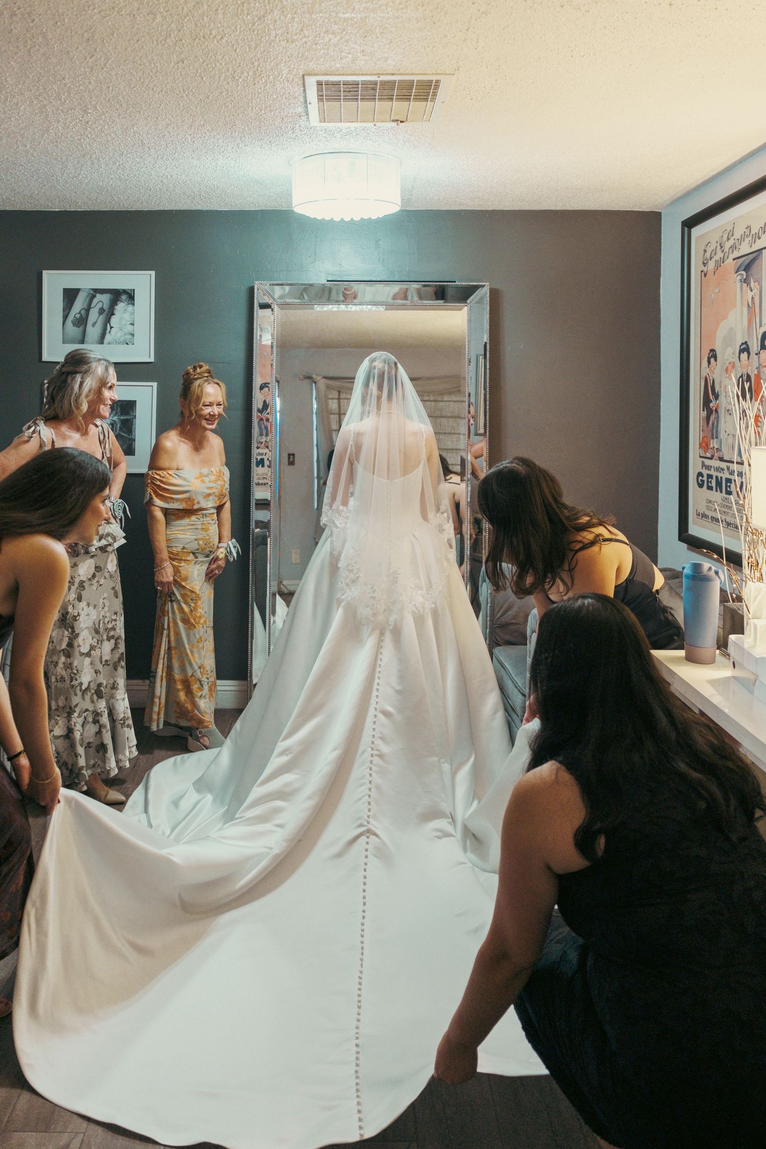 Bride in white gown and veil surrounded by bridesmaids adjusting her dress train before a large mirror.