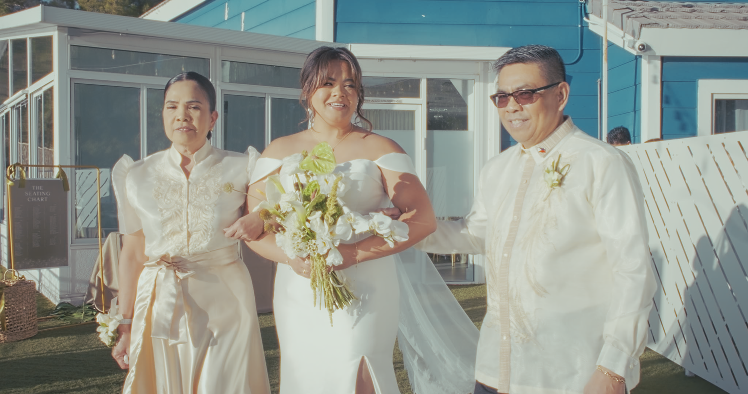 Bride in white off-shoulder gown poses with two family members in elegant white attire outdoors.