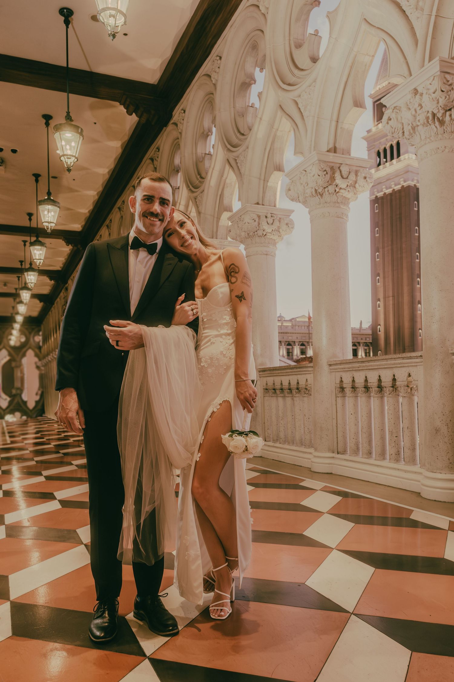 Smiling bride and groom pose in elegant columned corridor with ornate architecture and checkered floors.