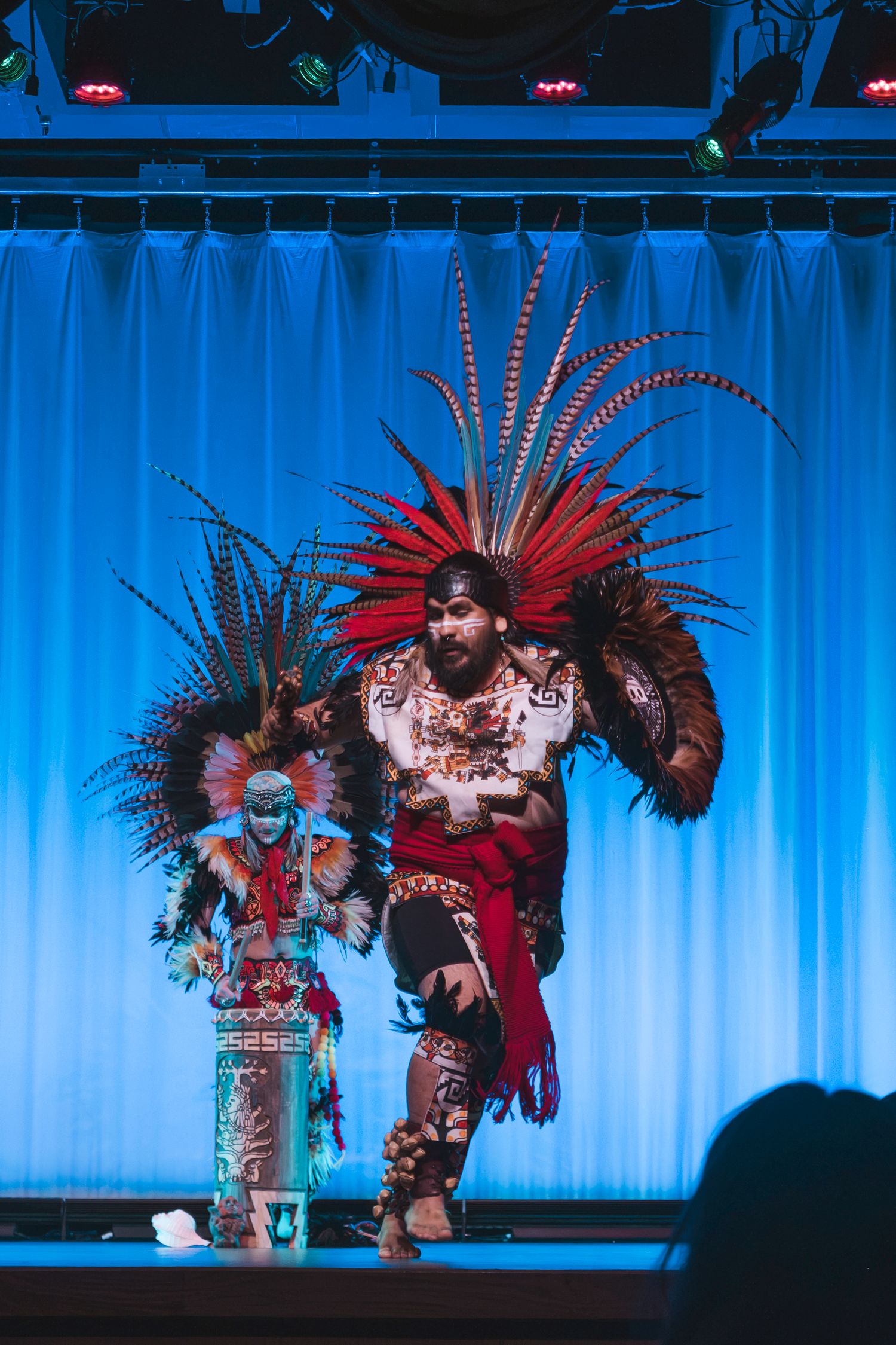 Two performers in vibrant Aztec ceremonial costumes with feathered headdresses dance on a blue-lit stage.