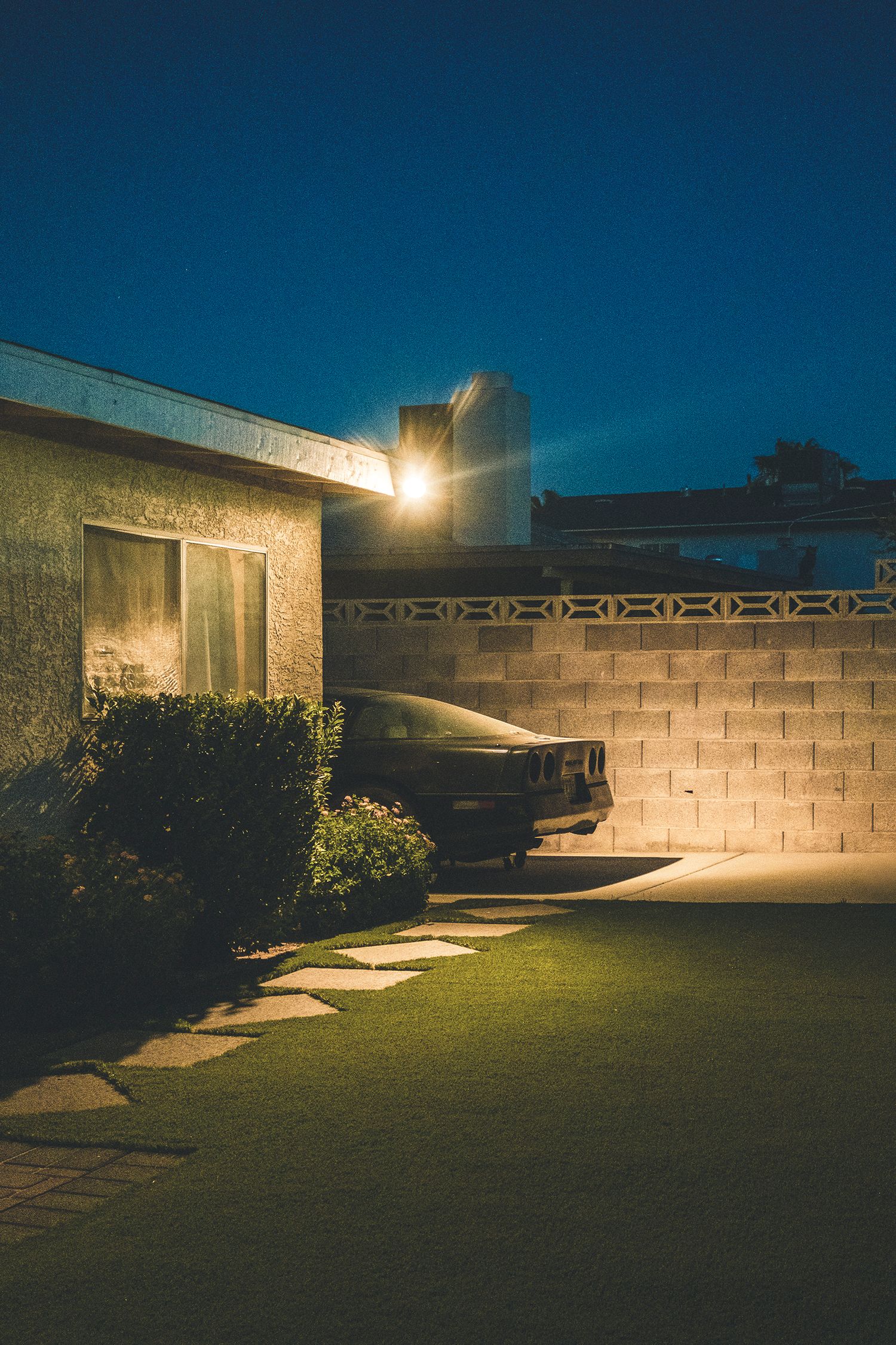 Vintage car parked beside a mid-century home at night, illuminated by a warm outdoor light with lush green lawn.
