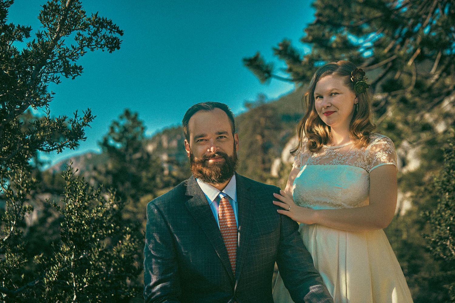 Couple in wedding attire posing outdoors with mountain trees and blue sky backdrop.