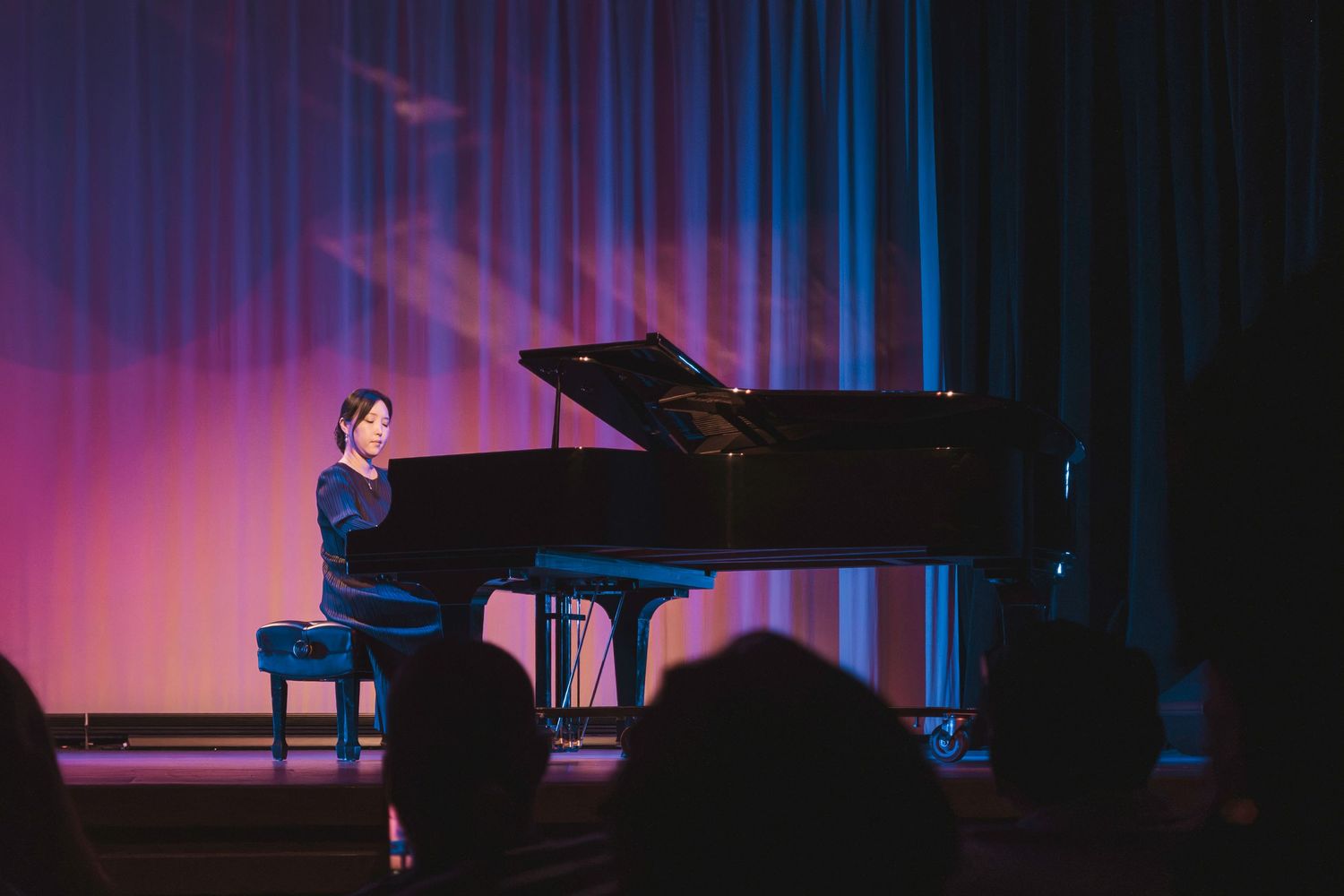 A pianist performs on a grand piano on a softly lit stage with pink and blue curtains backdrop.