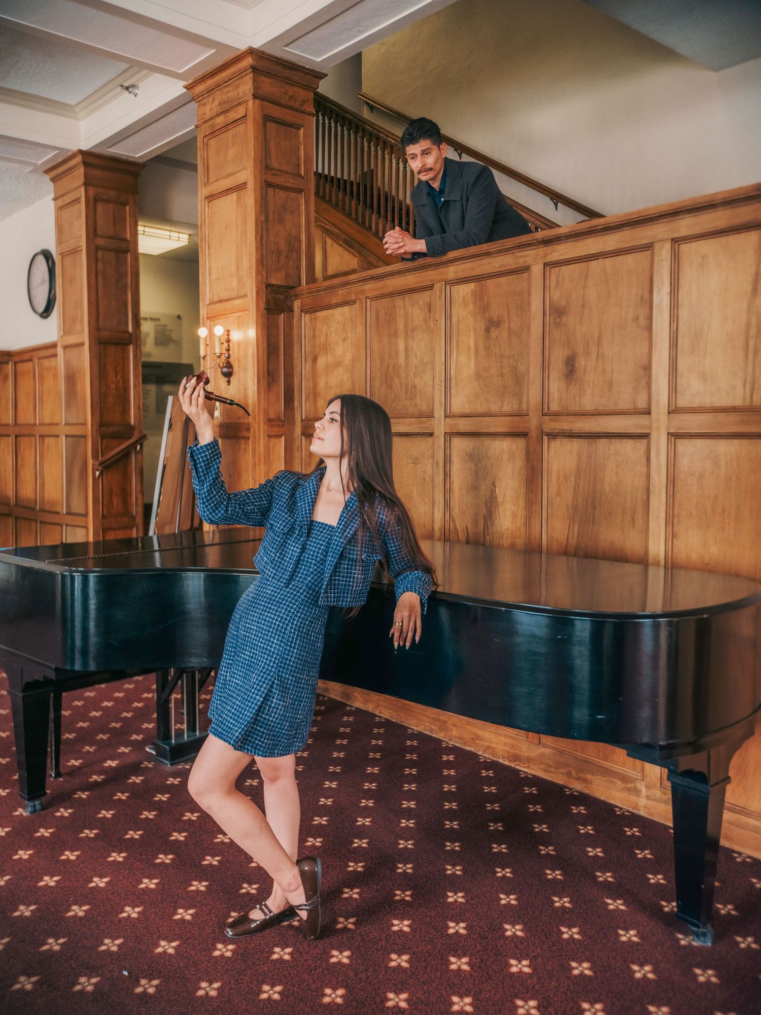Woman in blue suit taking selfie by grand piano while man watches from balcony in elegant wood-paneled room.