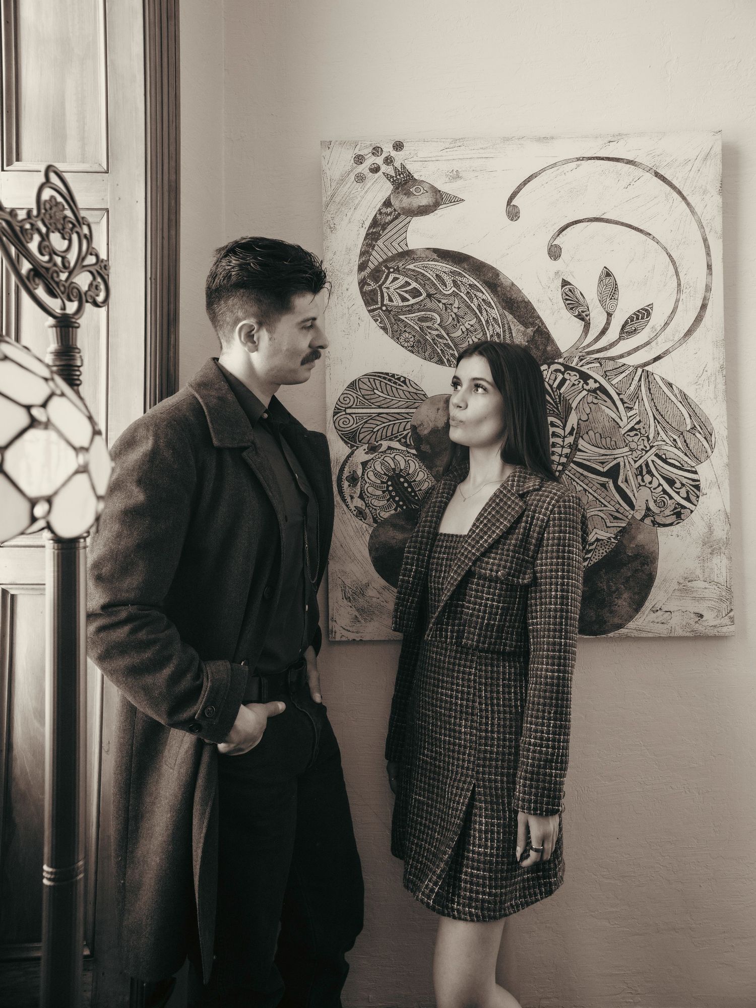 A couple in vintage-style outfits stands before a decorative peacock painting in a sepia-toned portrait photo.