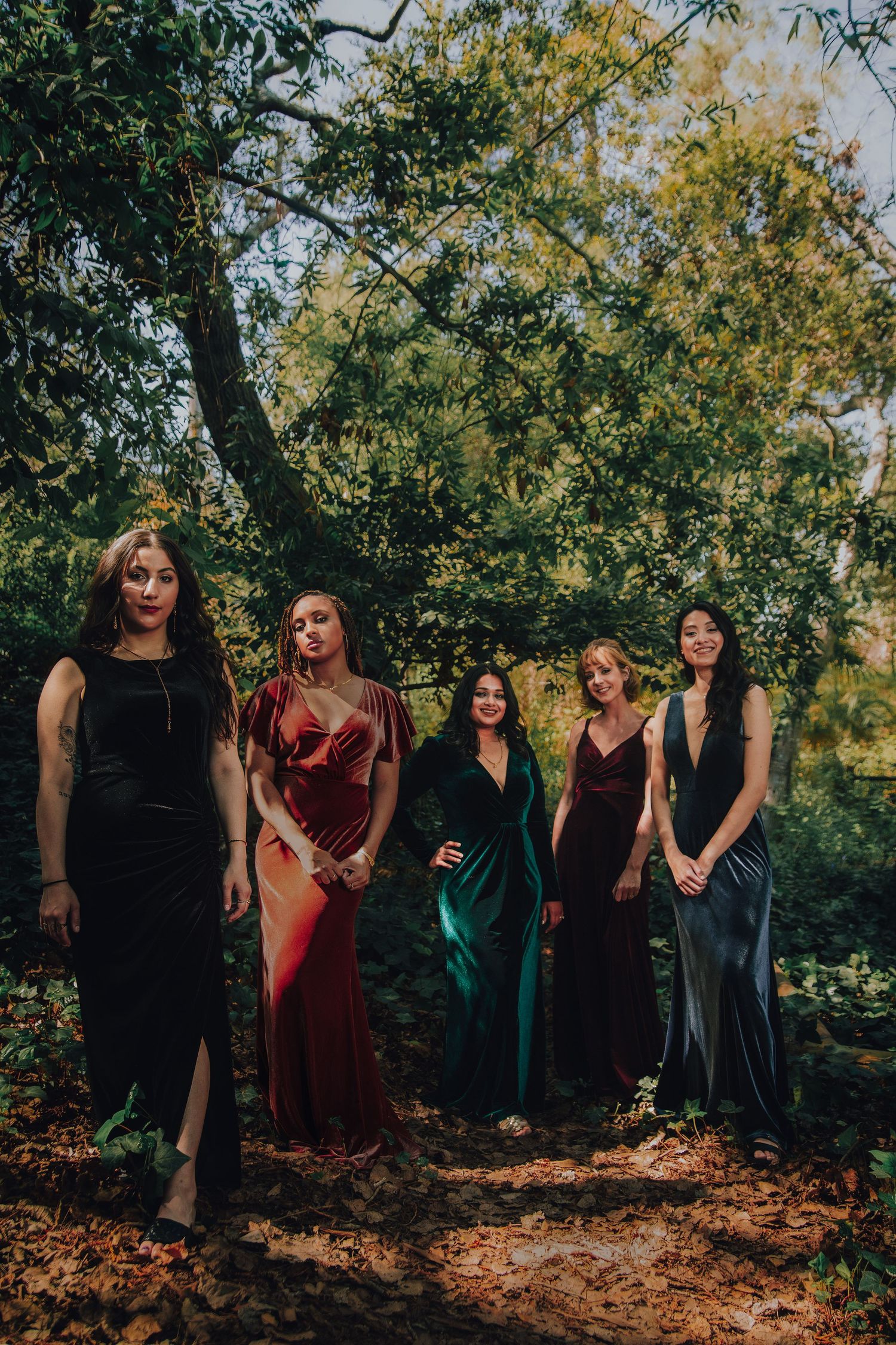 Five women in elegant velvet gowns pose outdoors among lush green trees in a forest setting.