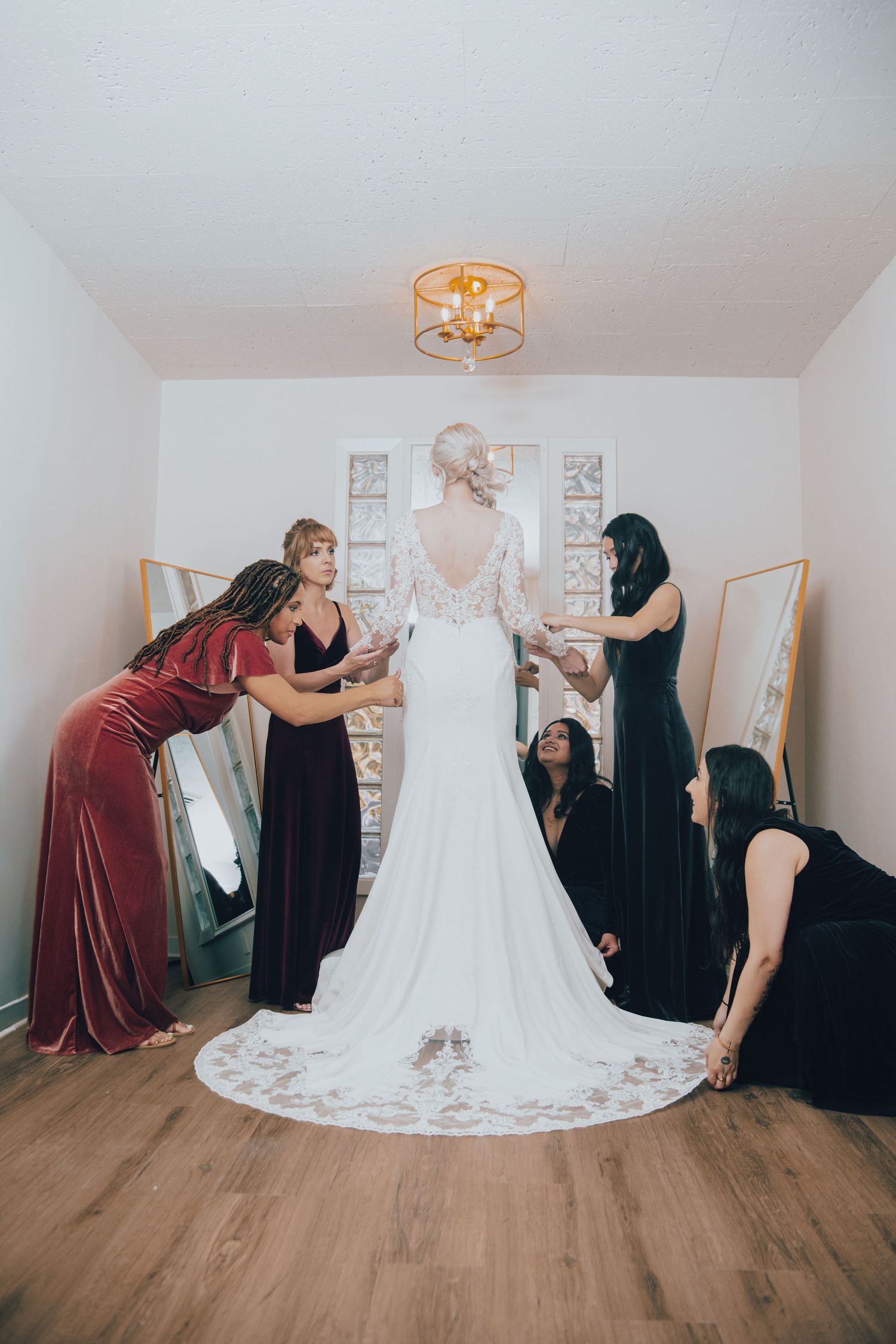 Bridesmaids in velvet gowns help a bride in a lace wedding dress with a train prepare in a bright dressing room.