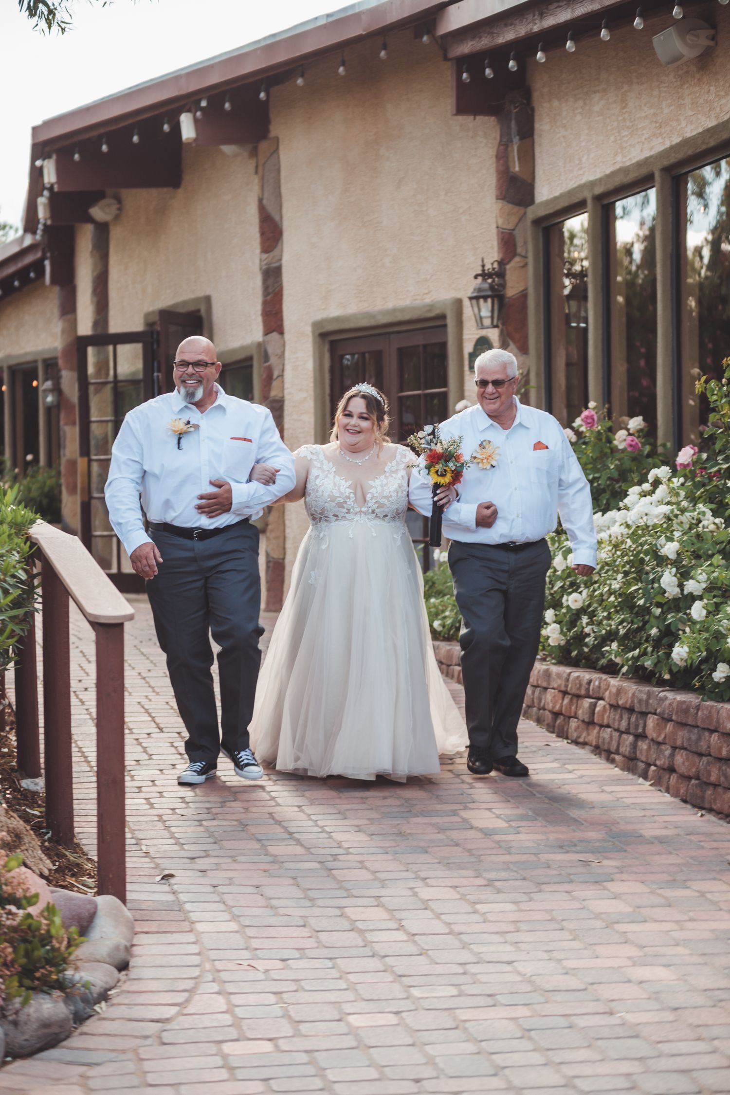 Bride in lace wedding gown walks down brick path flanked by two men in matching white shirts at outdoor venue.