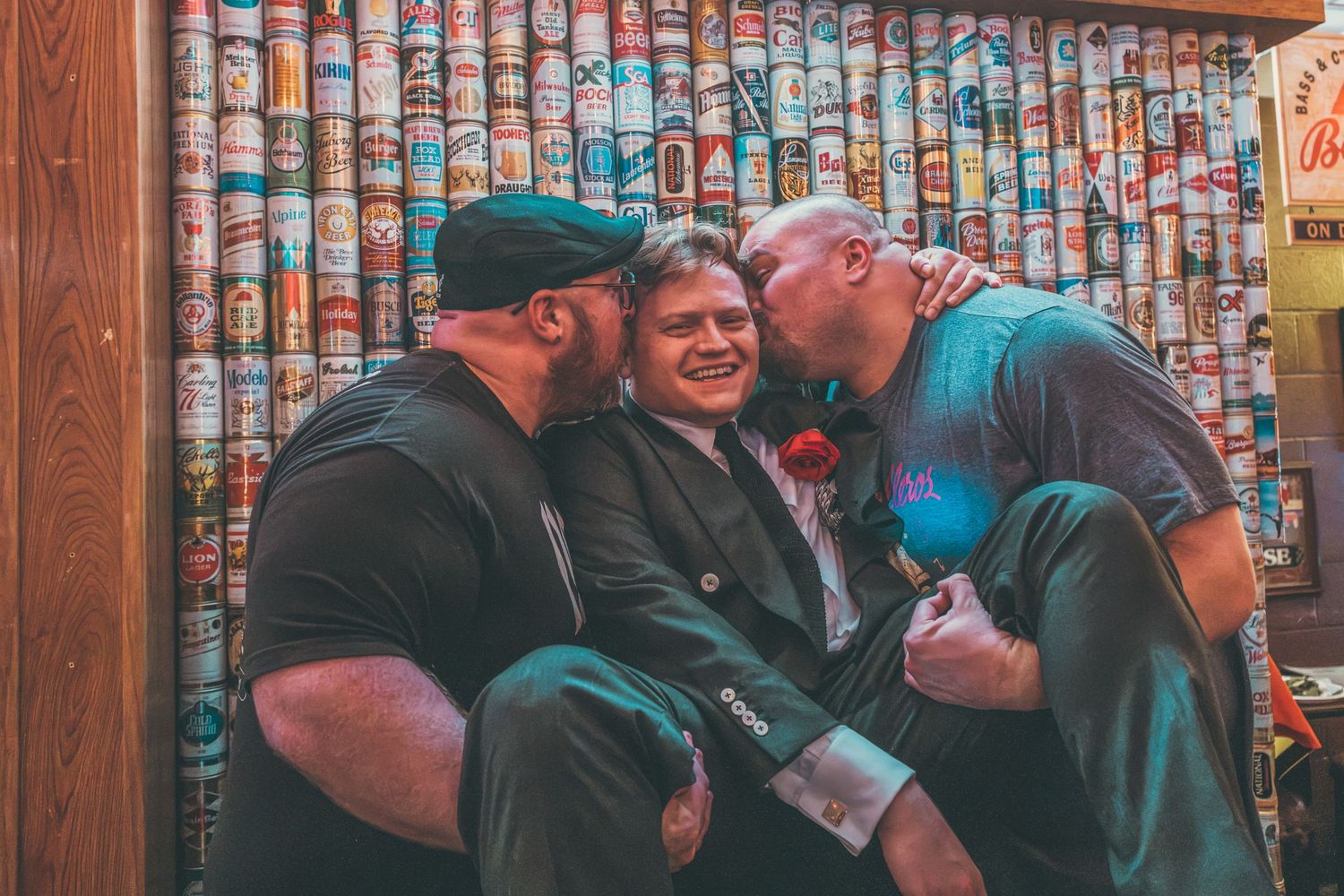 Three friends laugh together in front of a wall covered in colorful beer cans at a bar.