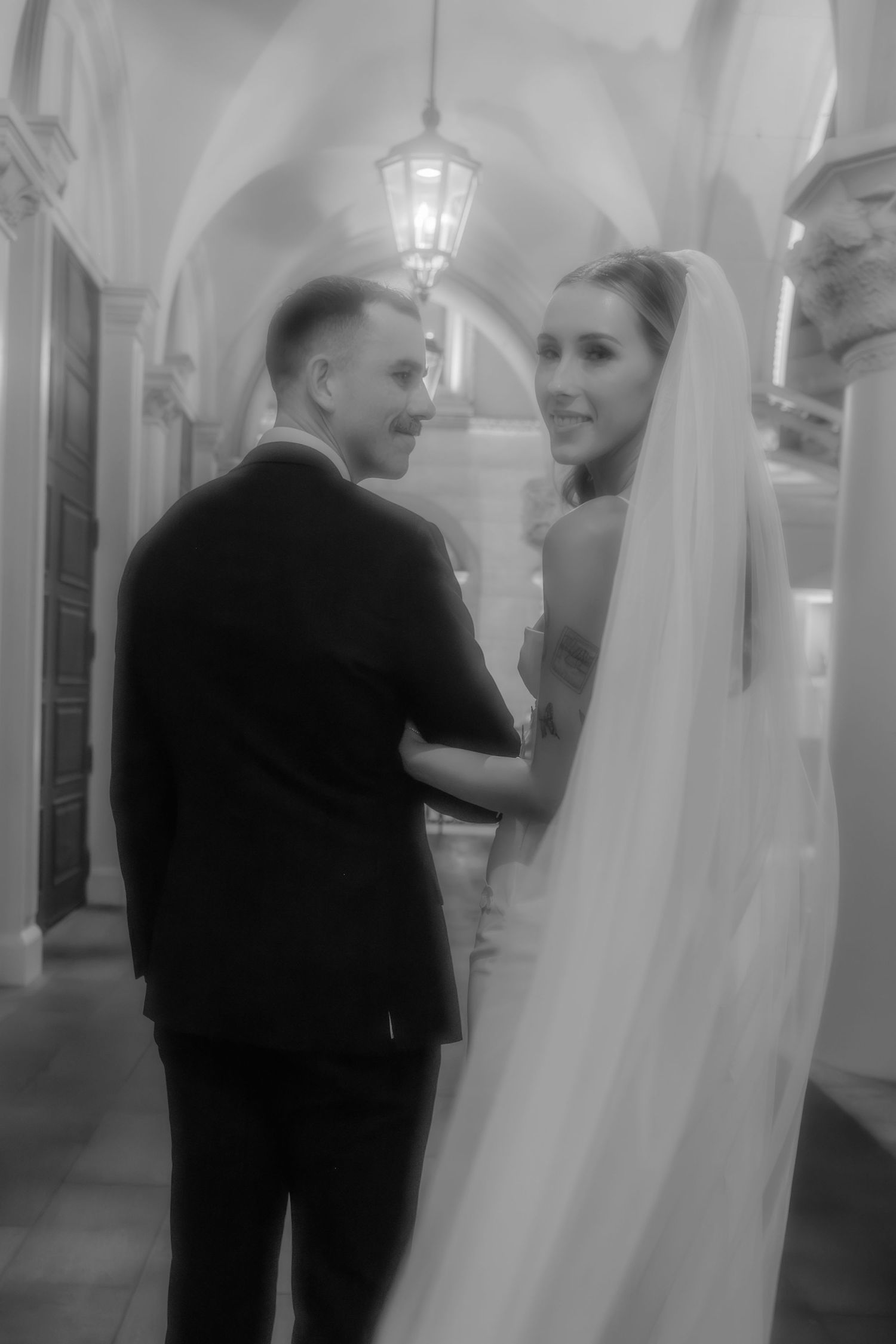Black and white photo of a bride and groom embracing in an elegant arched hallway.