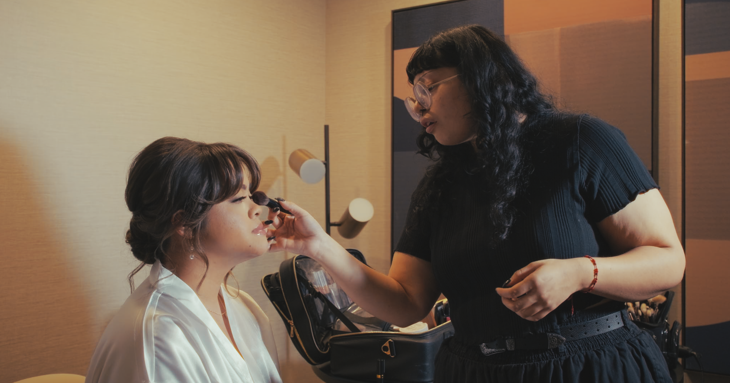 A makeup artist applies makeup to a bride during wedding preparations in a warmly lit room.