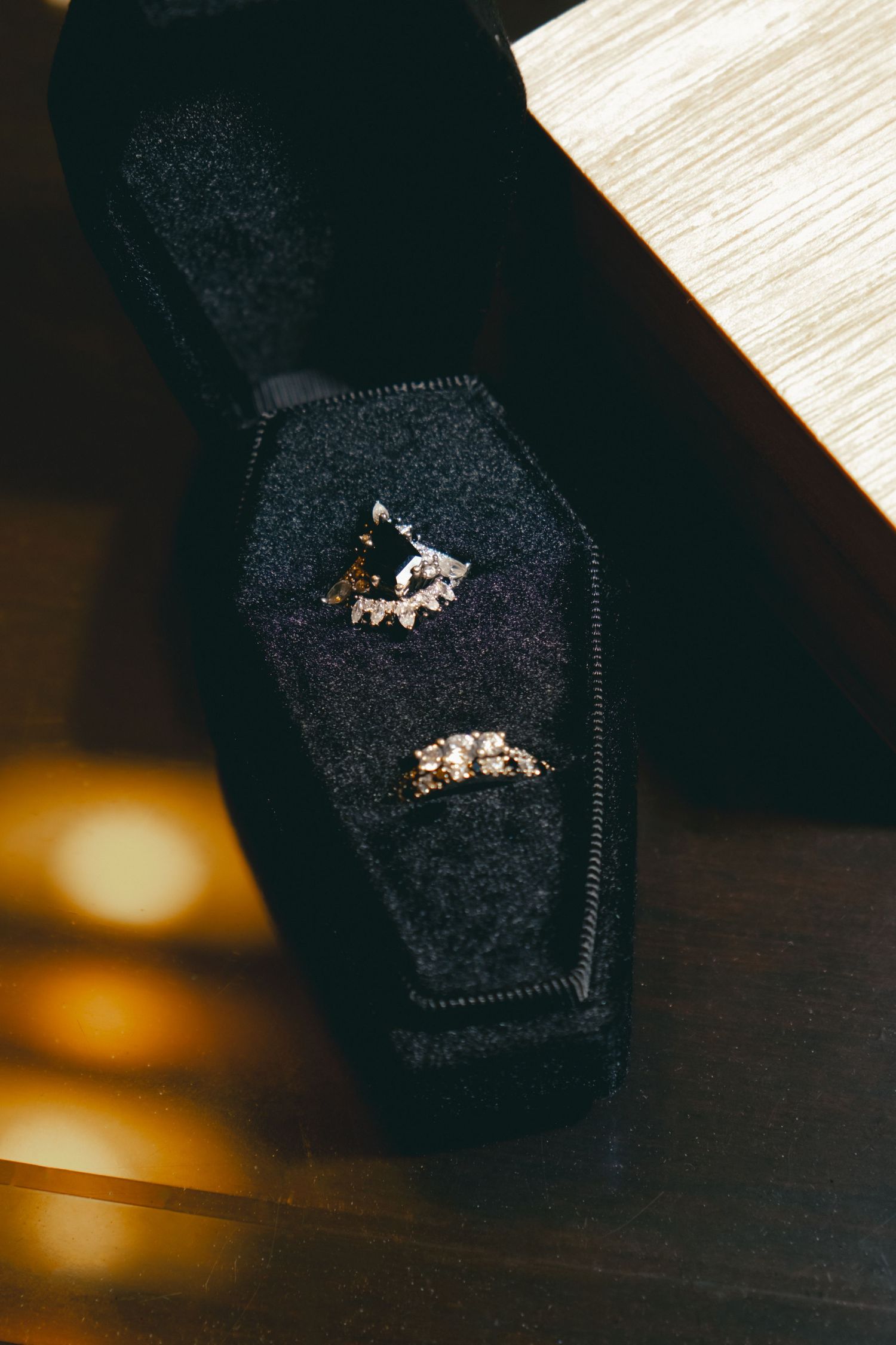Two diamond rings displayed in a black velvet coffin-shaped jewelry box on a wooden surface.