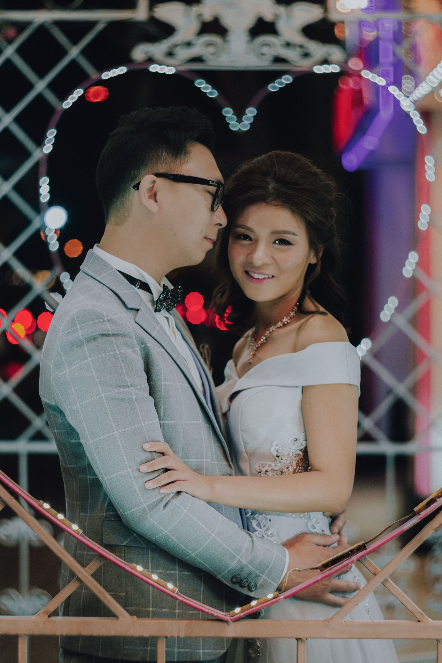 A couple in wedding attire embraces romantically, surrounded by colorful bokeh lights at night.