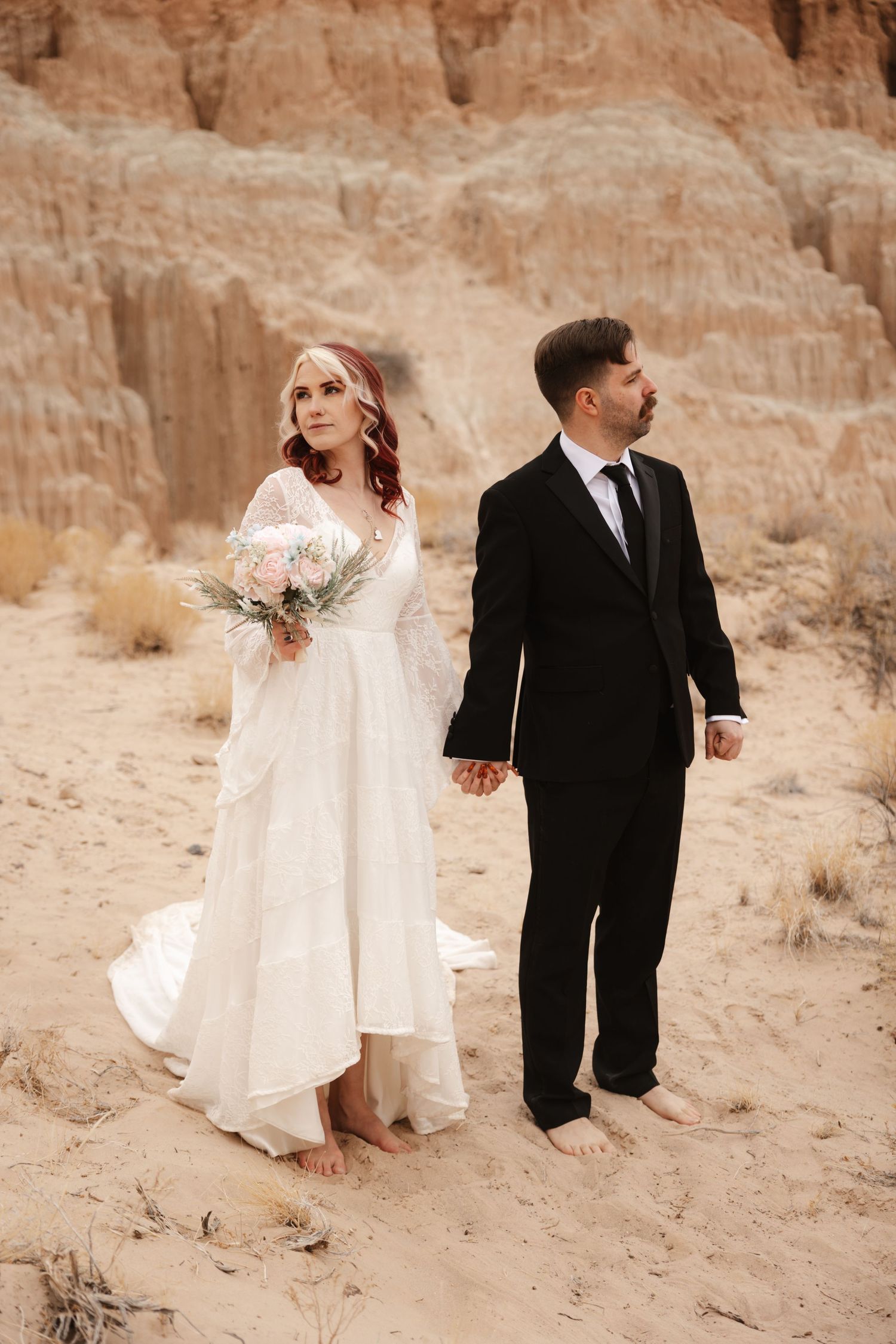 Barefoot bride holding bouquet and groom in black suit stand hand in hand in desert landscape.