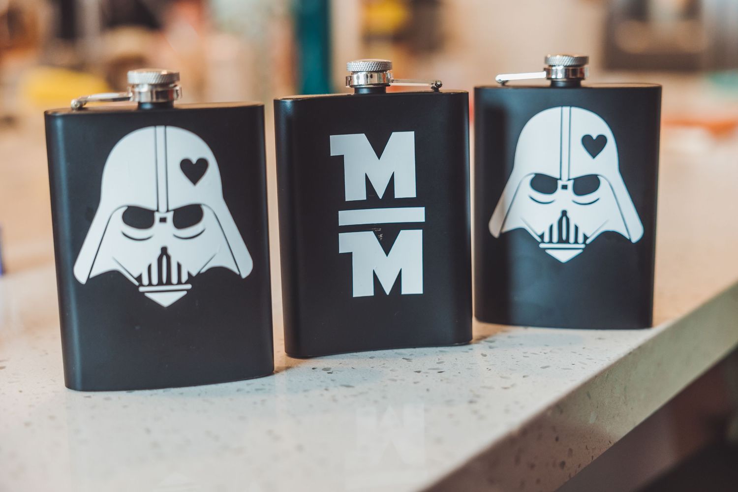 Three black hip flasks featuring Darth Vader helmet designs and monogram MM initials displayed on a surface.