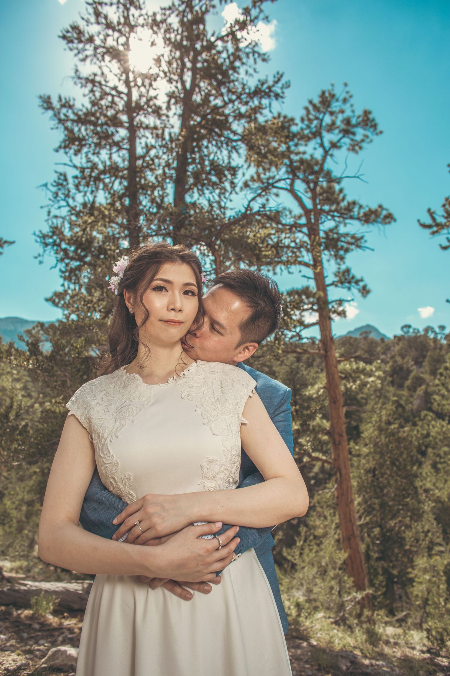 Couple embracing outdoors among pine trees, groom kissing bride's cheek in white dress during mountain wedding shoot.