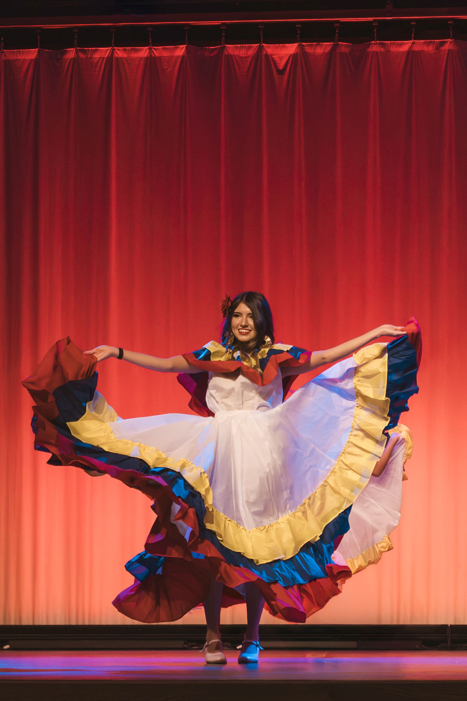 Woman performing traditional Latin folk dance on stage in colorful flowing dress with red curtain backdrop.