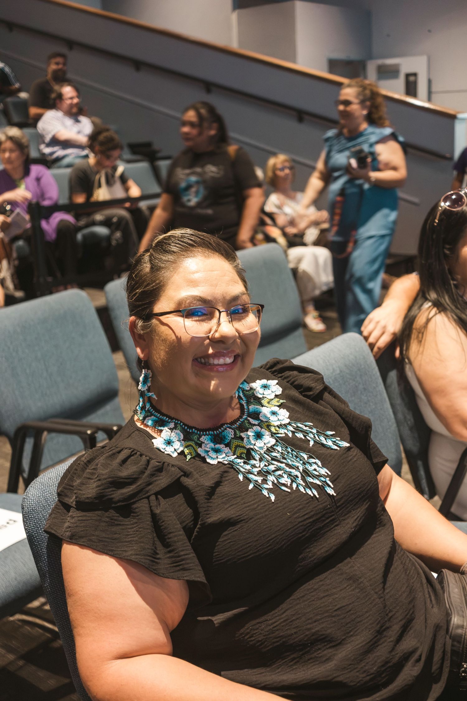 Woman in black floral dress smiling in auditorium seating, wearing glasses, surrounded by diverse group of attendees.