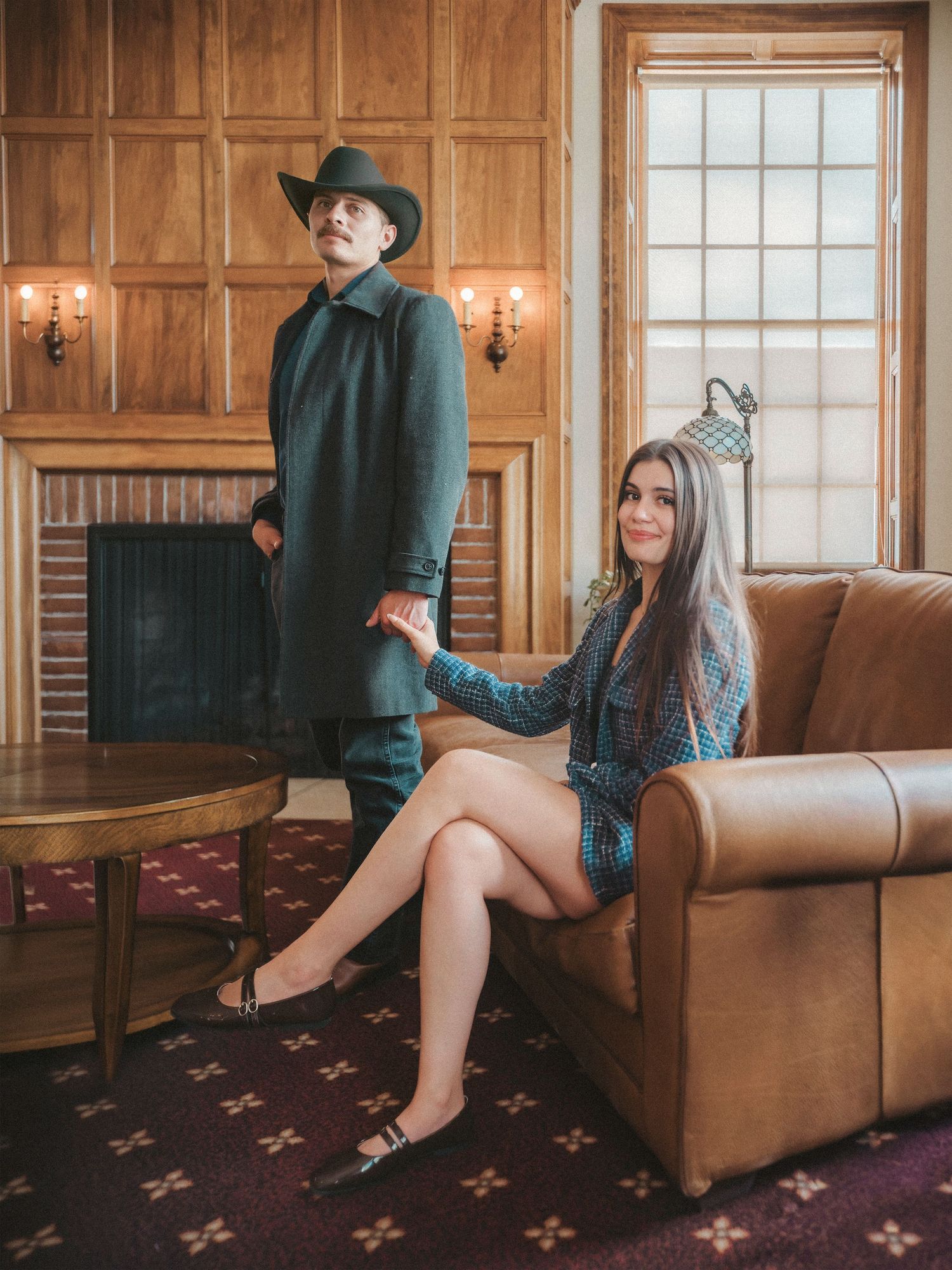 A couple poses in a wood-paneled room, man in cowboy hat and coat, woman seated in leather chair.