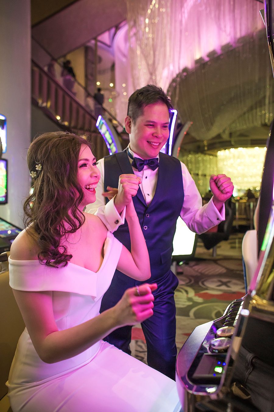 Excited newlywed couple celebrating a casino win at slot machines, bride in white dress and groom in tuxedo vest.