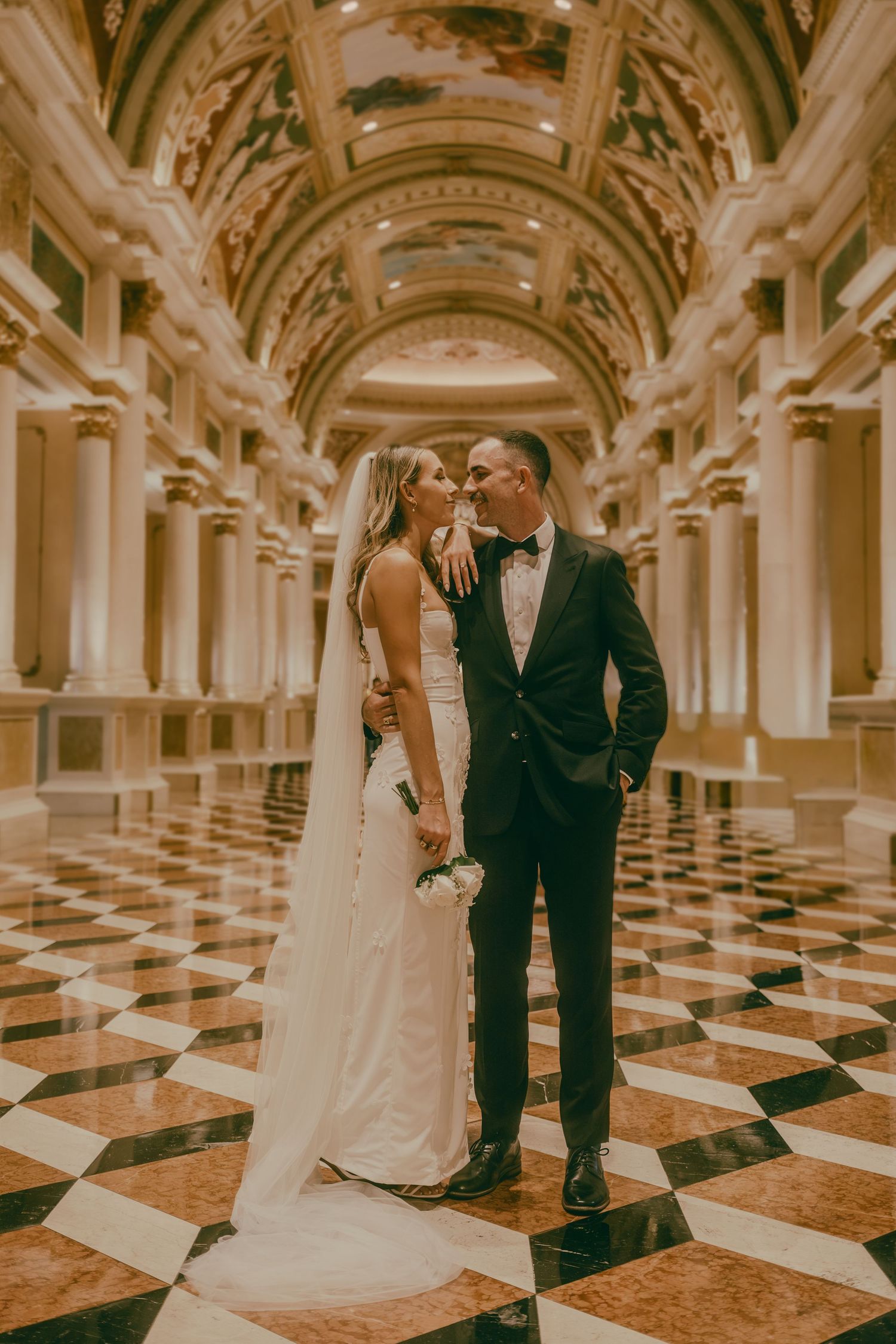 Bride and groom posing in opulent marble-floored ballroom with ornate ceiling.